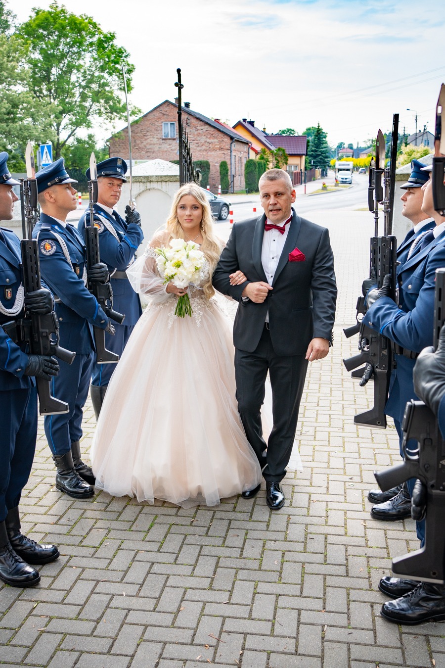 Para młoda przechodzi pod szpalerem żołnierzy w galowych mundurach z karabinami podczas uroczystości ślubnej.