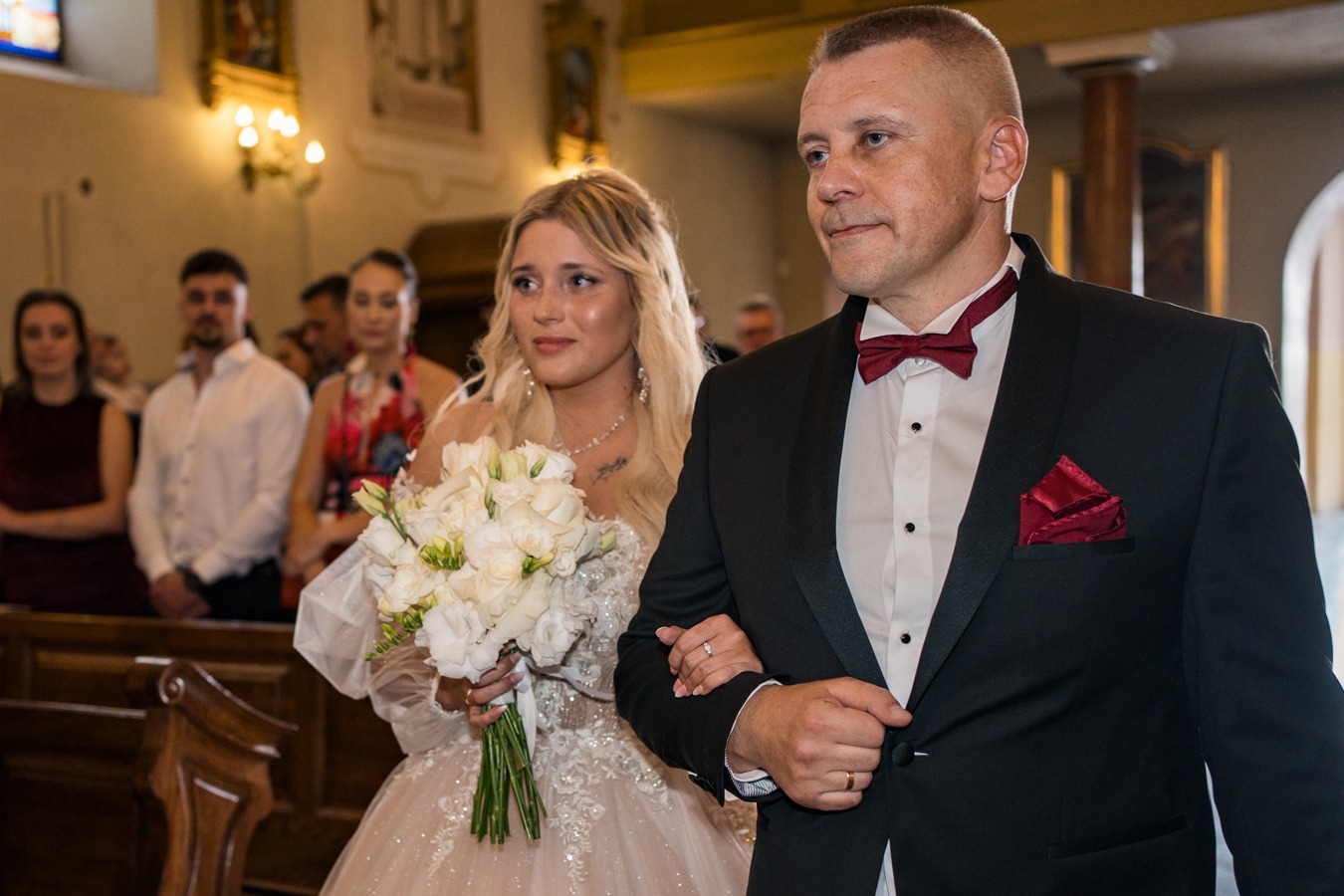 Para młoda w kościele: panna młoda z bukietem białych kwiatów w sukni z koronką, pan młody w czarnym smokingu z bordową muchą i poszetką, w tle goście.