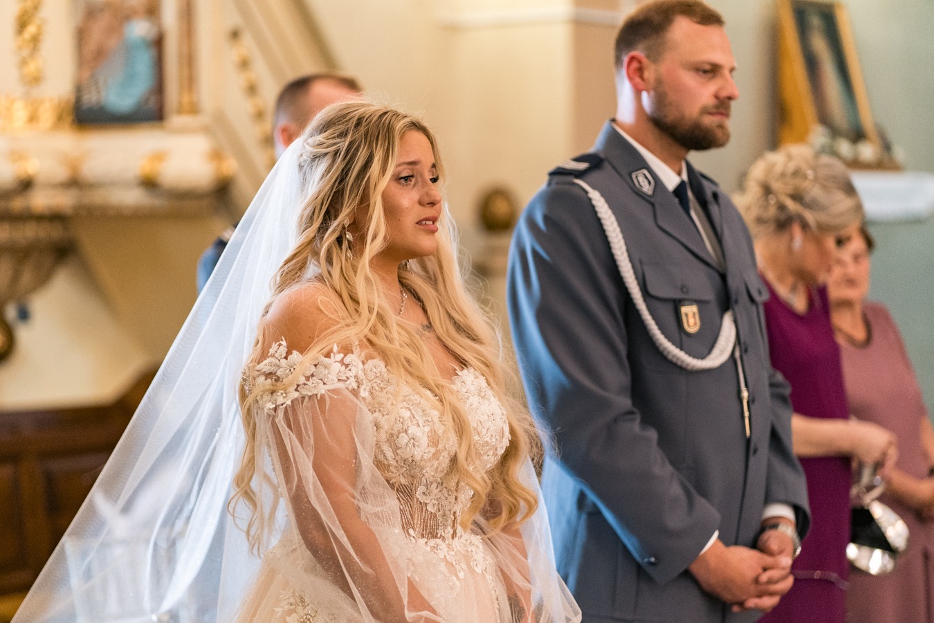 Panna młoda w koronkowej sukni ślubnej i długim welonie, obok mężczyzna w mundurze galowym, w tle wnętrze kościoła.