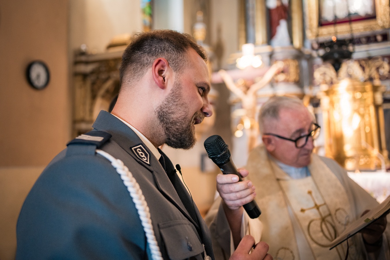 Mężczyzna w mundurze policyjnym przemawia do mikrofonu podczas uroczystości kościelnej, w tle ksiądz z księgą.