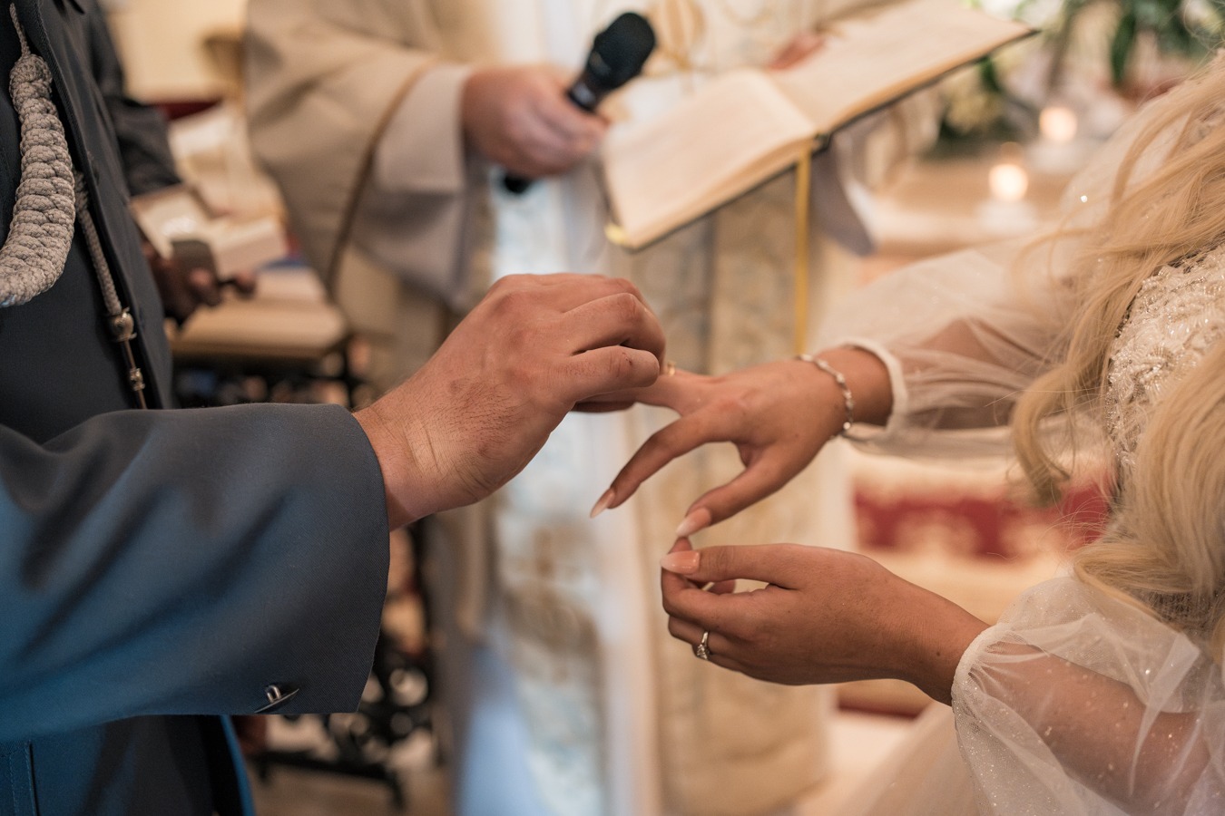 Wymiana obrączek podczas ceremonii ślubnej, panna młoda w zdobionej sukni, pan młody w eleganckim garniturze, ksiądz z otwartą księgą i mikrofonem w tle.
