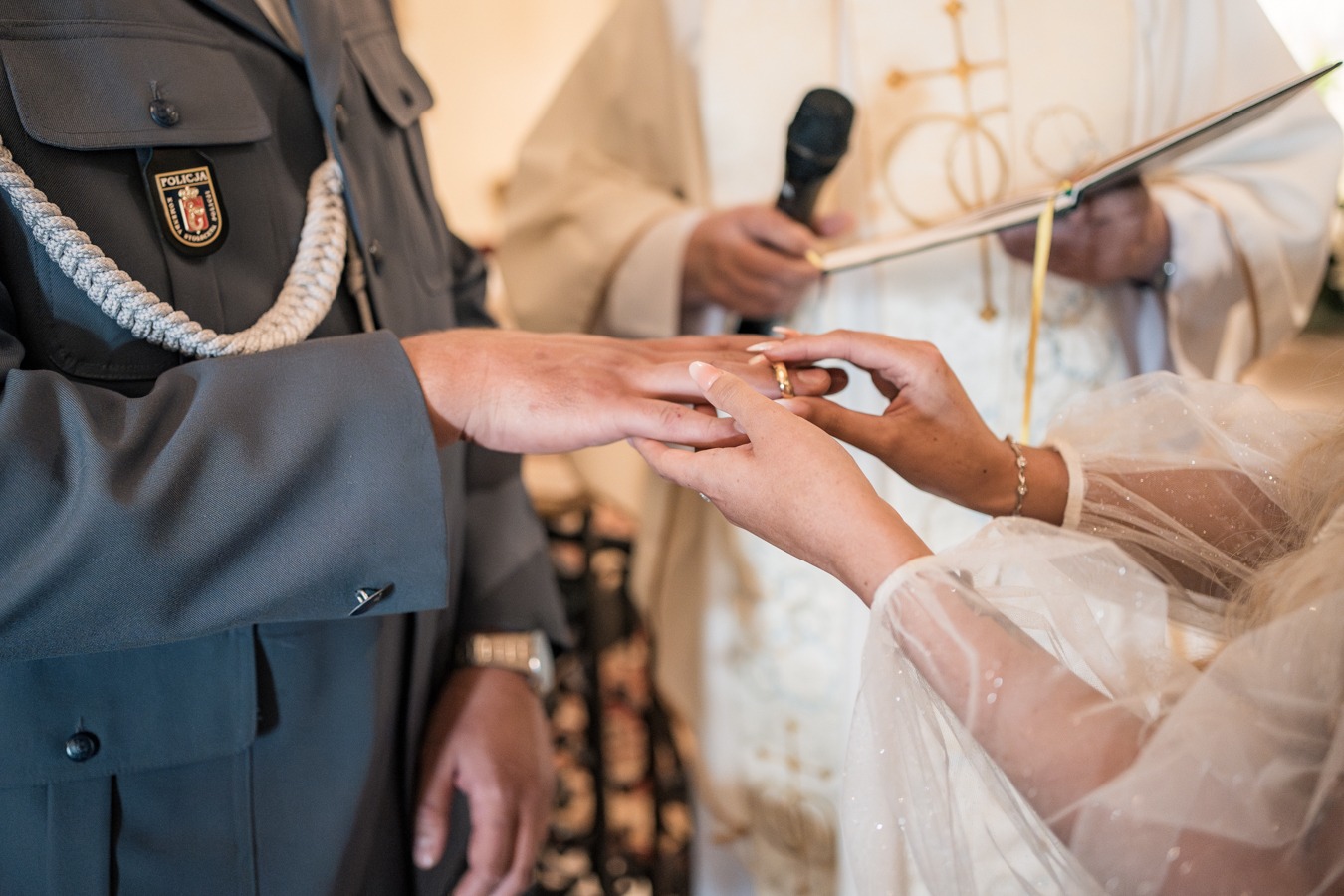Wymiana obrączek podczas ceremonii ślubnej, mężczyzna w mundurze policyjnym, kobieta w białej sukni, w tle ksiądz z mikrofonem i księgą.