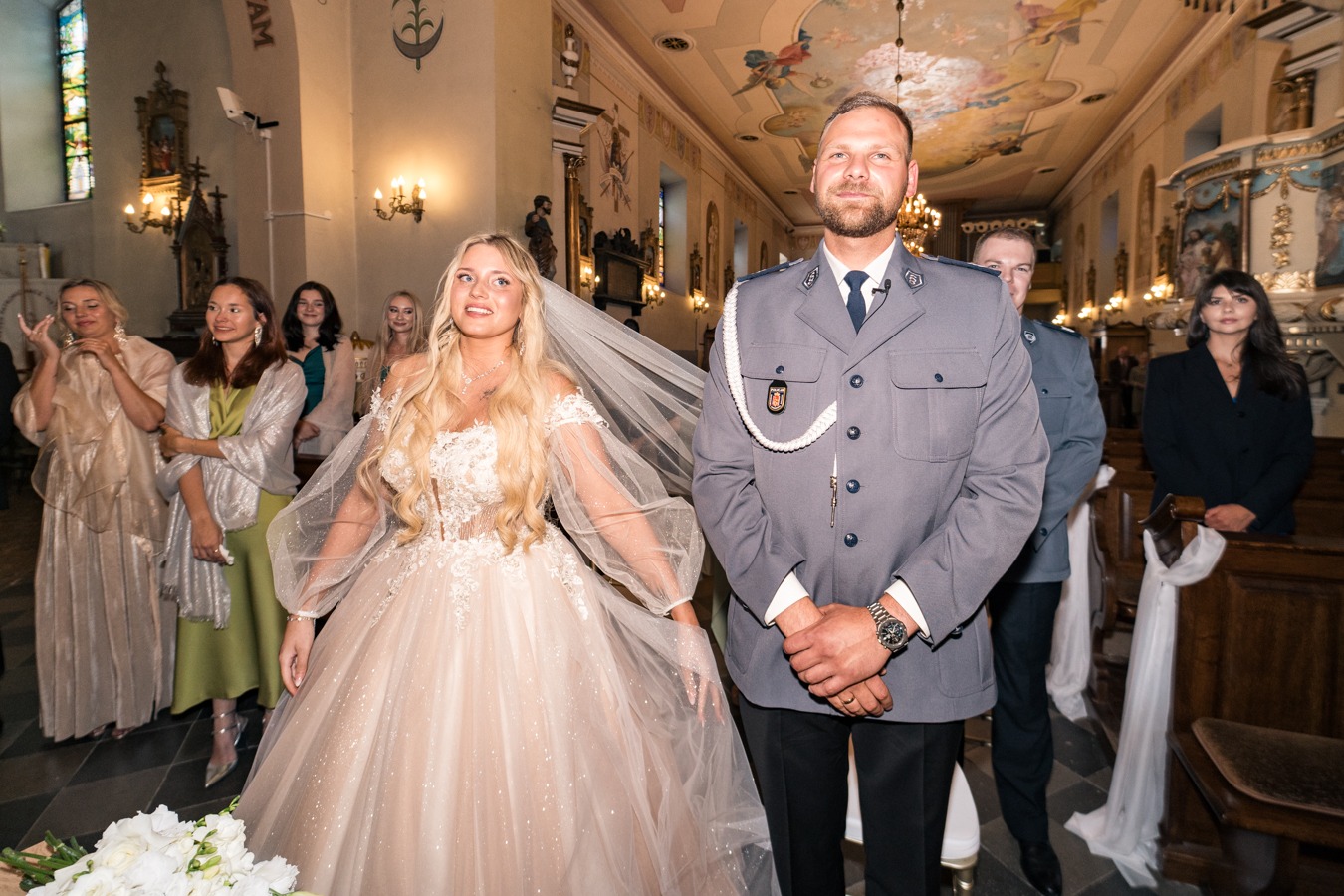 Panna młoda w sukni ślubnej z tiulowymi rękawami i welonem obok mężczyzny w szarym mundurze policyjnym w kościele, goście w tle.