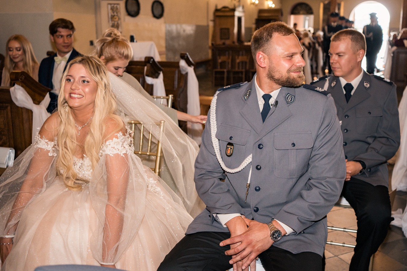 Uśmiechnięta panna młoda w sukni z koronkowymi rękawami i welonem siedzi obok pana młodego w mundurze policyjnym podczas ceremonii ślubnej w kościele.