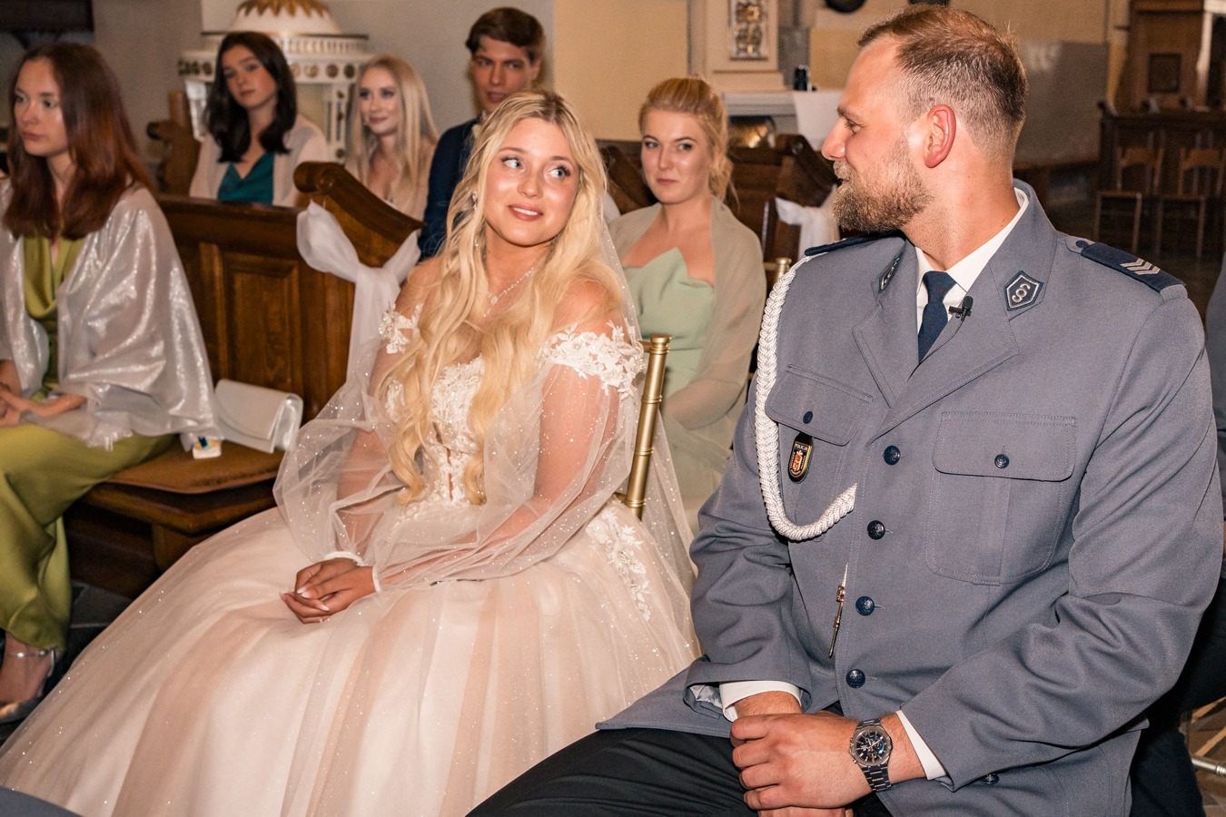 Panna młoda w sukni ślubnej z długimi rękawami, siedząca obok mężczyzny w mundurze policyjnym podczas ceremonii w kościele, z gośćmi w tle.