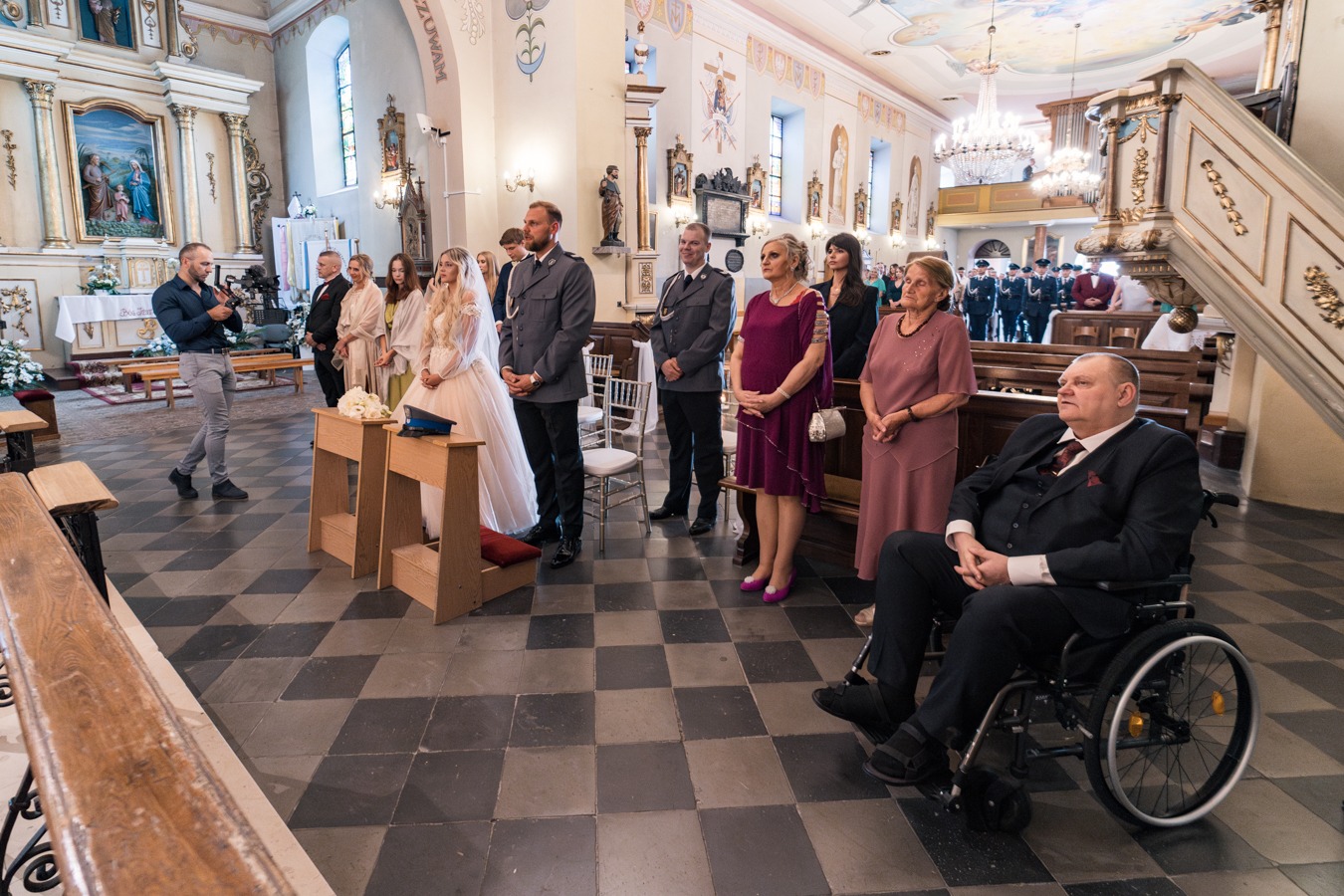 Para młoda w kościele podczas ceremonii ślubnej, goście w tle, mężczyzna robiący zdjęcia, posadzka w szachownicę.