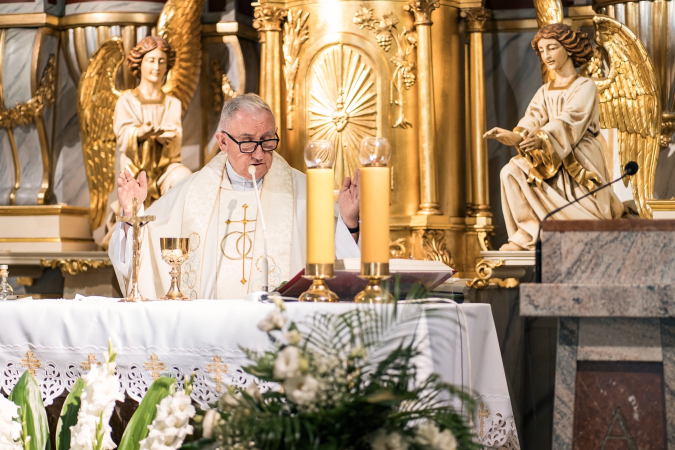 Ksiądz w okularach podczas ceremonii chrztu przy ołtarzu z aniołami, świecami i kwiatami.