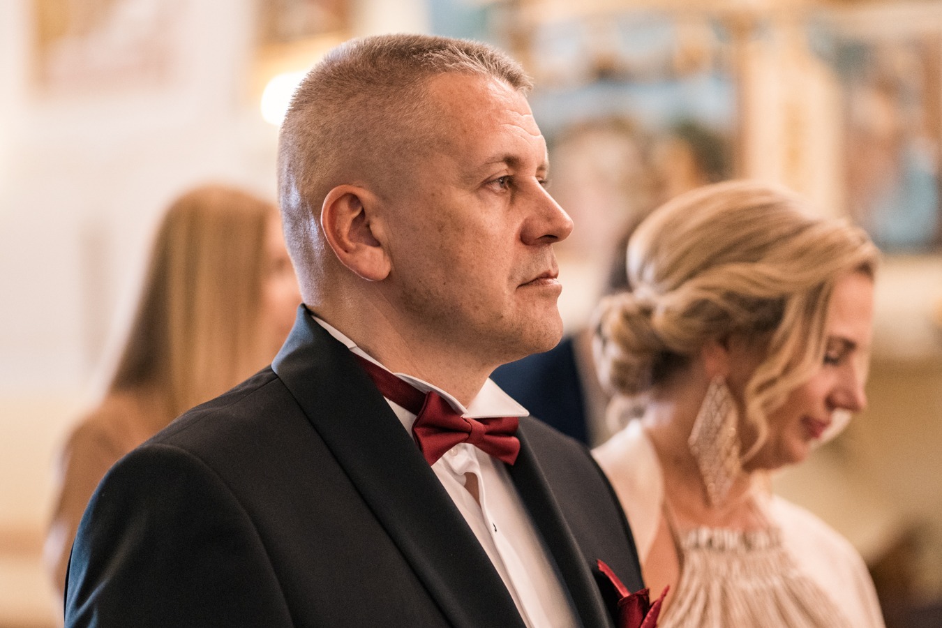 Mężczyzna w czarnym smokingu z bordową muchą i poszetką, stojący obok kobiety z upiętymi blond włosami i dużymi kolczykami w kształcie rombów, w jasnym wnętrzu kościoła.