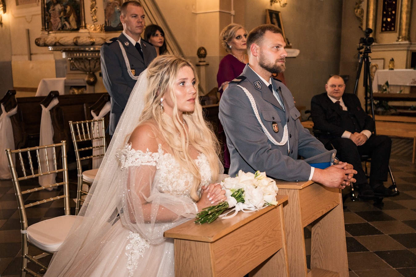 Panna młoda w koronkowej sukni ślubnej i welonie, obok pan młody w mundurze galowym, podczas ceremonii w kościele, z bukietem białych róż na klęczniku.