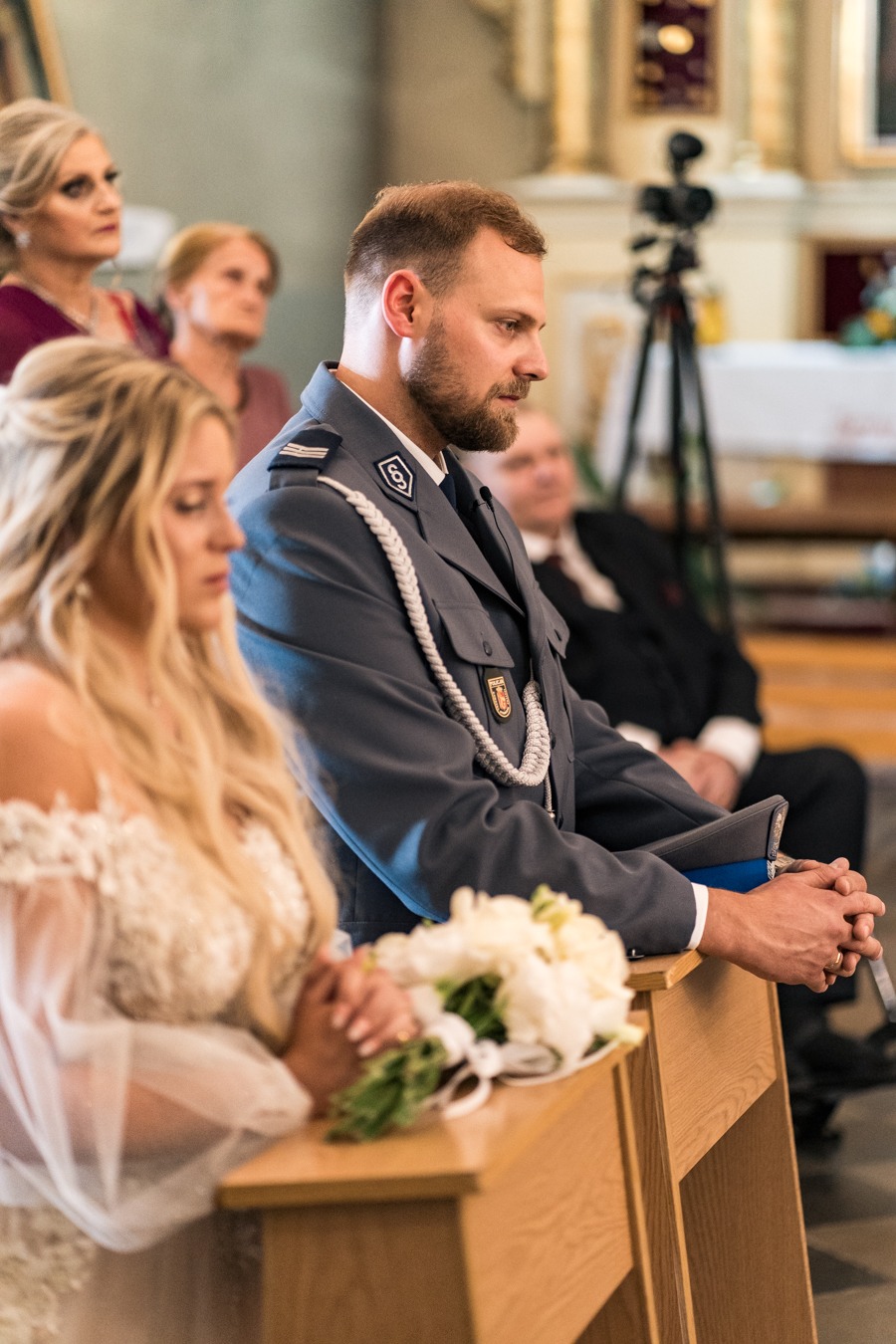 Para młoda podczas ceremonii ślubnej w kościele: panna młoda w koronkowej sukni, pan młody w mundurze galowym policji, bukiet białych kwiatów na klęczniku.