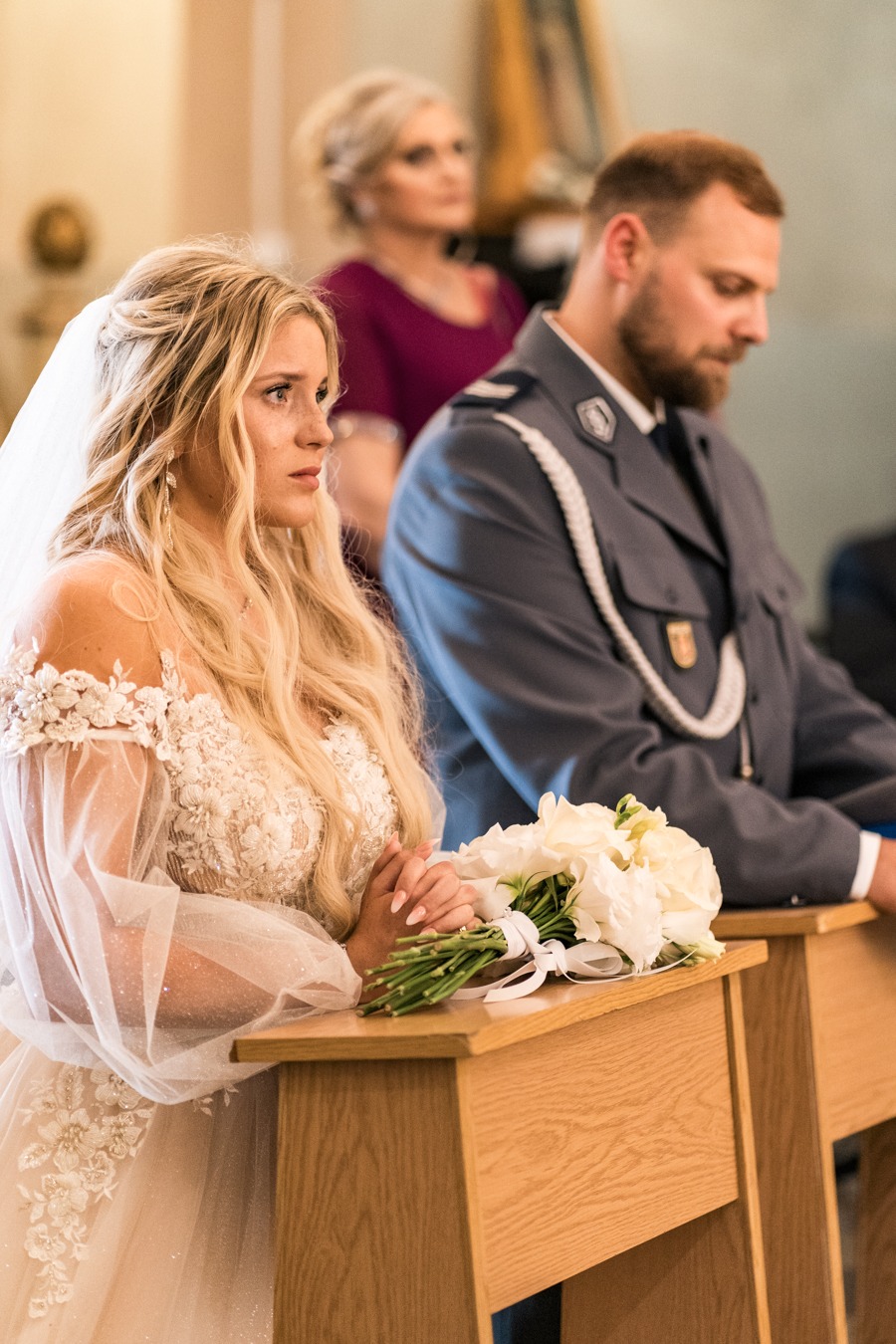 Panna młoda w koronkowej sukni i welonie, z bukietem białych kwiatów, klęczy w kościele obok mężczyzny w mundurze galowym.