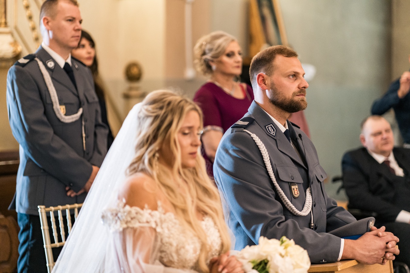 Panna młoda w białej sukni i welonie siedzi obok mężczyzny w mundurze galowym policji podczas ceremonii ślubnej w kościele.