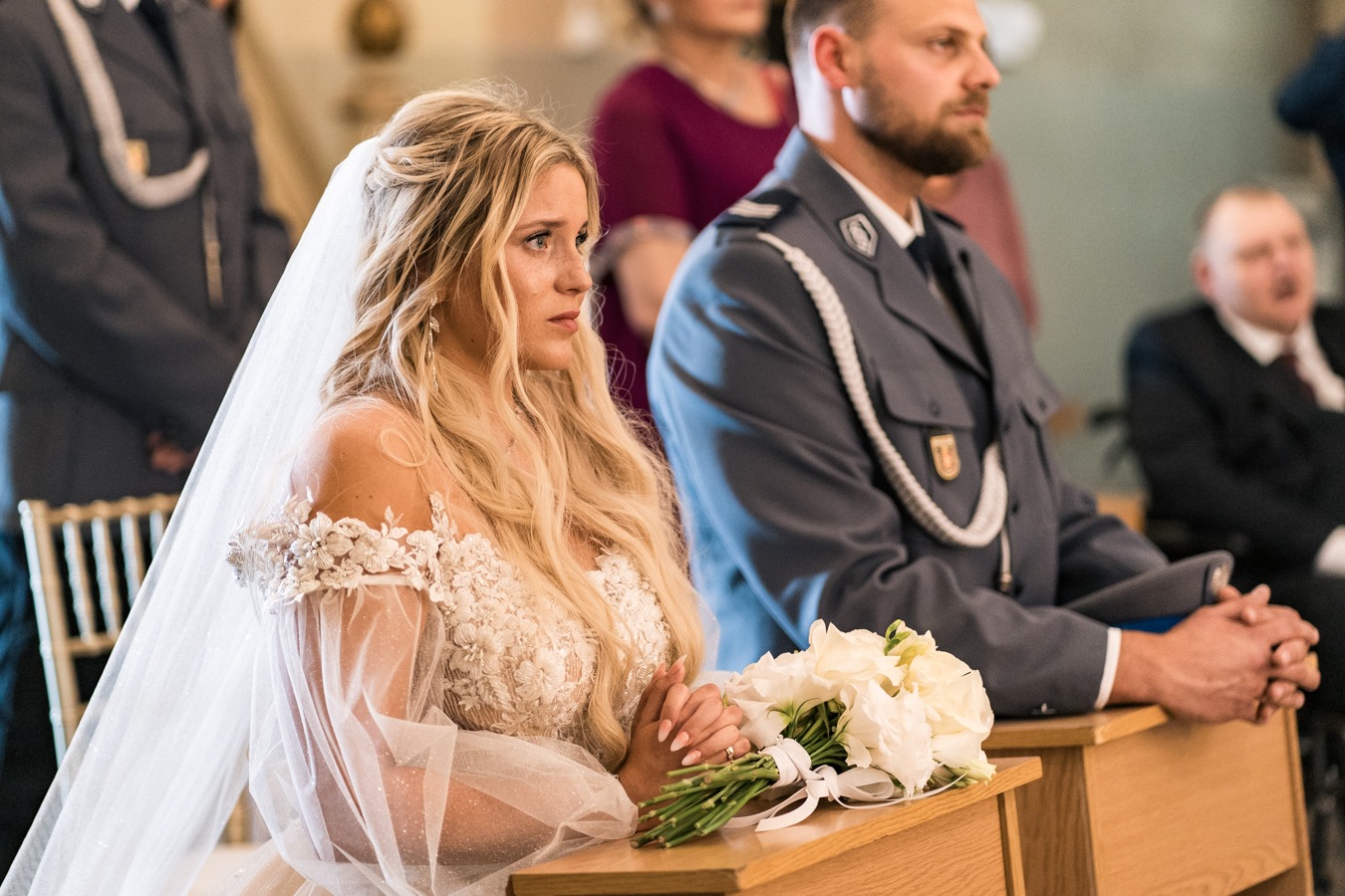 Panna młoda w koronkowej sukni i welonie, z zamyśloną miną, siedzi obok pana młodego w mundurze policyjnym podczas ceremonii ślubnej w kościele.
