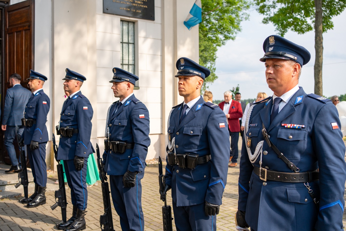 Uroczysta zmiana warty: szereg umundurowanych funkcjonariuszy w granatowych uniformach i czapkach, trzymających karabiny, stoi na tle budynku z tablicą pamiątkową i flagą.