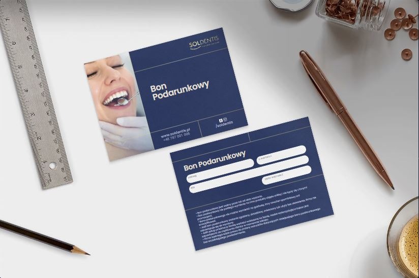 Dwa granatowe bony podarunkowe z logo Soldentis leżące na białym biurku obok metalowej linijki, ołówka, złotego długopisu i szklanki z kawą.