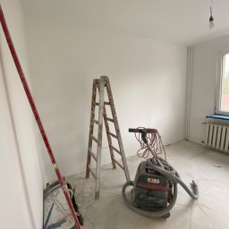JANUSZ KOŹMIC - Pokój po szpachlowaniu i malowaniu, widoczna drabina, poziomica oparta o ścianę, szlifierka do gładzi z odkurzaczem, ściany pomalowane na biało, okno zabezpieczone folią.
