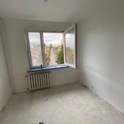 JANUSZ KOŹMIC - Pokój w trakcie przygotowania do malowania: goła żarówka, odrapana podłoga, zaklejone okno z widokiem na ulicę, grzejnik i sprzęt do szlifowania podłogi.