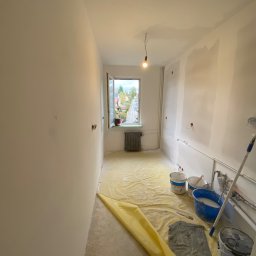 JANUSZ KOŹMIC - Wnętrze pokoju w trakcie przygotowania do malowania: ściany po szpachlowaniu, podłoga zabezpieczona folią, widoczne wiadra z farbą, wałek malarski i wisząca żarówka.