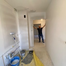 JANUSZ KOŹMIC - Korytarz w trakcie prac wykończeniowych: szpachlowane ściany, przygotowane narzędzia malarskie (wałki, pędzle, wiadra z farbą), zabezpieczona podłoga folią, mężczyzna w tle z uniesionymi rękami.