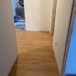 JANUSZ KOŹMIC - Układanie paneli podłogowych imitujących drewno w trakcie remontu łazienki, widoczne niedokończone ściany i przygotowane materiały w tle.