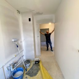 JANUSZ KOŹMIC - Wnętrze korytarza w trakcie prac wykończeniowych; widoczne ślady szpachlowania na ścianach, wiadro z farbą, wałek malarski oraz mężczyzna w oddali, opierający się o futrynę.