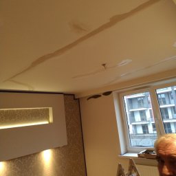 JANUSZ KOŹMIC - Pokój w trakcie prac wykończeniowych: widoczne ślady po szpachlowaniu na suficie, drabina oparta o ścianę przy oknie, fragment twarzy osoby w prawym dolnym rogu kadru.