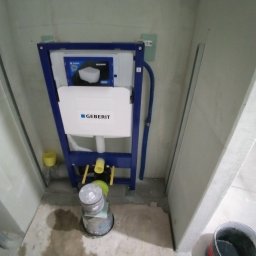 JANUSZ KOŹMIC - Stelaż podtynkowy Geberit Sigma zamontowany w surowej łazience, widoczne rury, żółte elementy instalacji i puszki z zaprawą na podłodze.