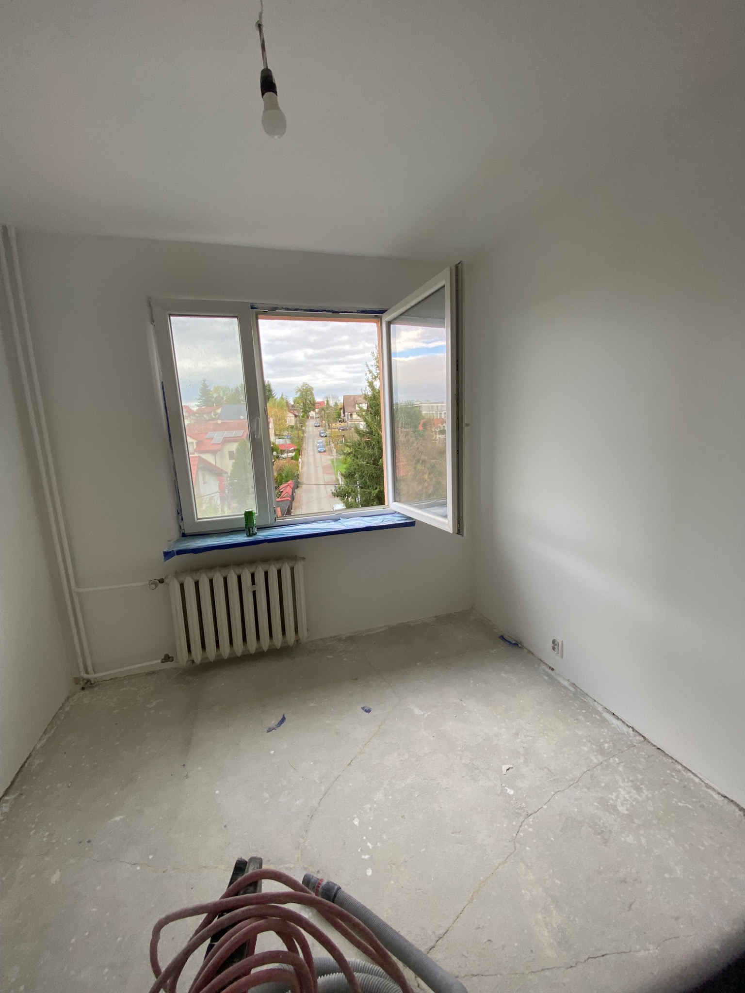Pokój w trakcie przygotowania do malowania: goła żarówka, odrapana podłoga, zaklejone okno z widokiem na ulicę, grzejnik i sprzęt do szlifowania podłogi.