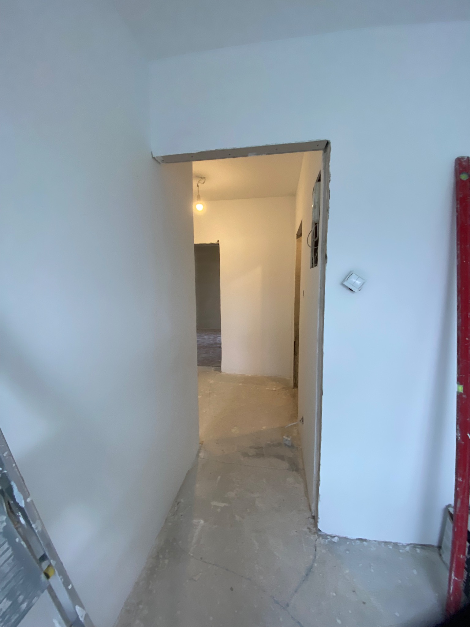 Korytarz w trakcie przygotowania do malowania, widoczne świeżo wyszpachlowane ściany, surowy beton na podłodze i wystający przełącznik elektryczny.