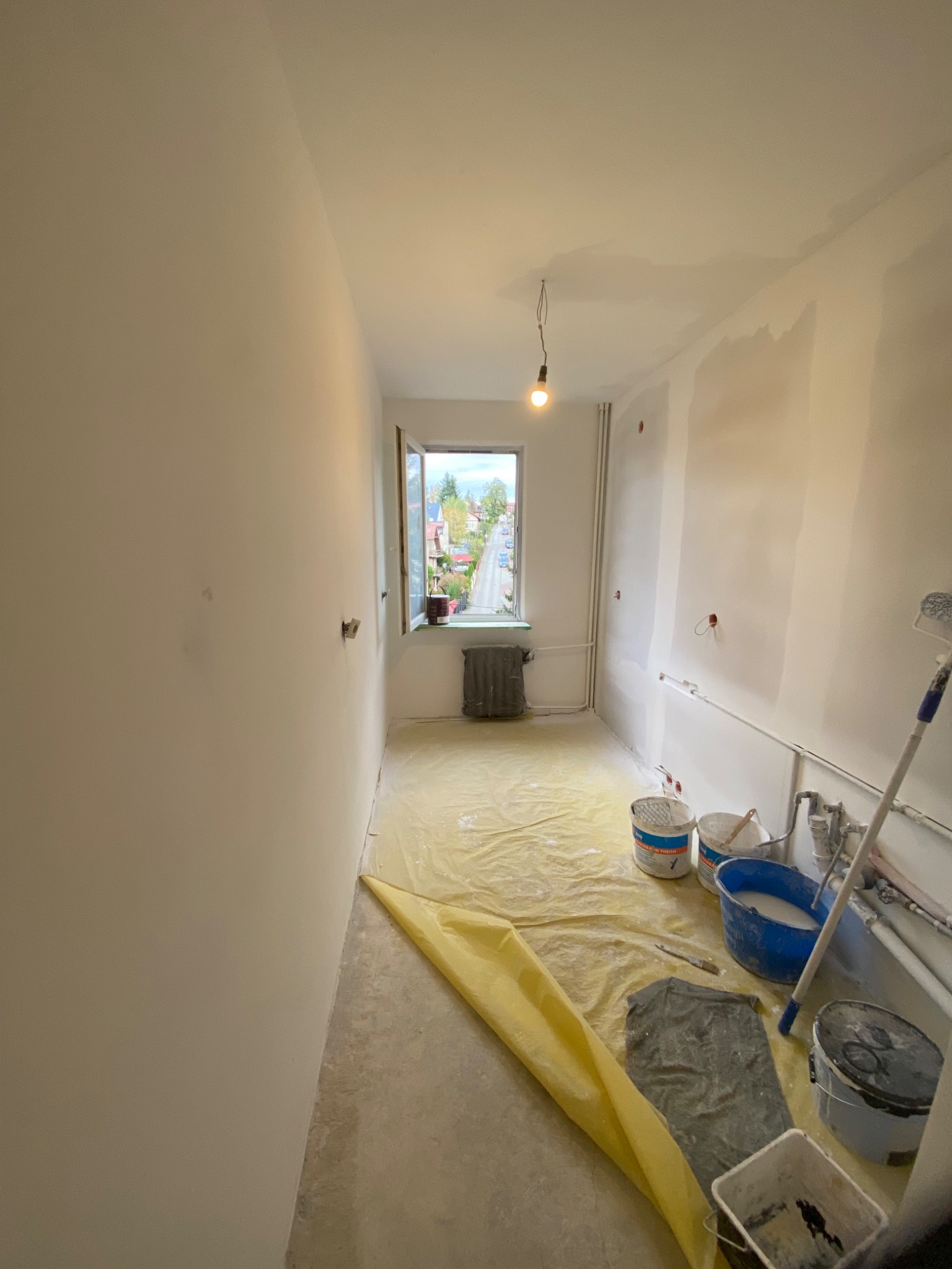 Wnętrze pokoju w trakcie przygotowania do malowania: ściany po szpachlowaniu, podłoga zabezpieczona folią, widoczne wiadra z farbą, wałek malarski i wisząca żarówka.