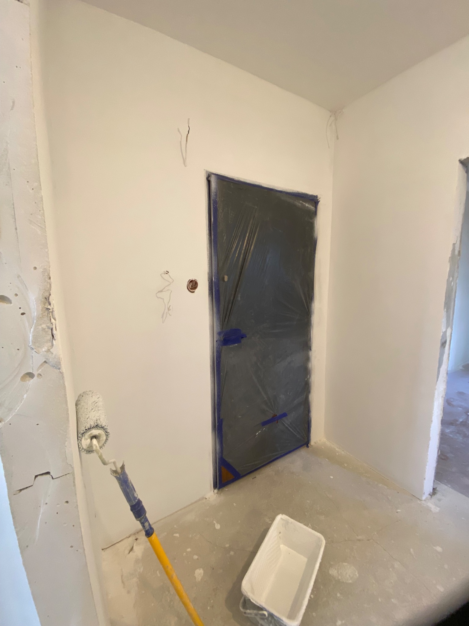 Pokój w trakcie odświeżania: ściany po pierwszym malowaniu na biało, zabezpieczone folią drzwi, wałek malarski oparty o ścianę, kuweta malarska na podłodze.