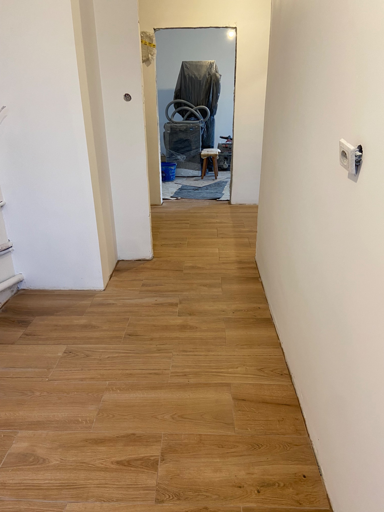Korytarz z nową, drewnopodobną podłogą z płytek, widoczna puszka elektryczna bez osprzętu i pomieszczenie w trakcie prac wykończeniowych w tle.