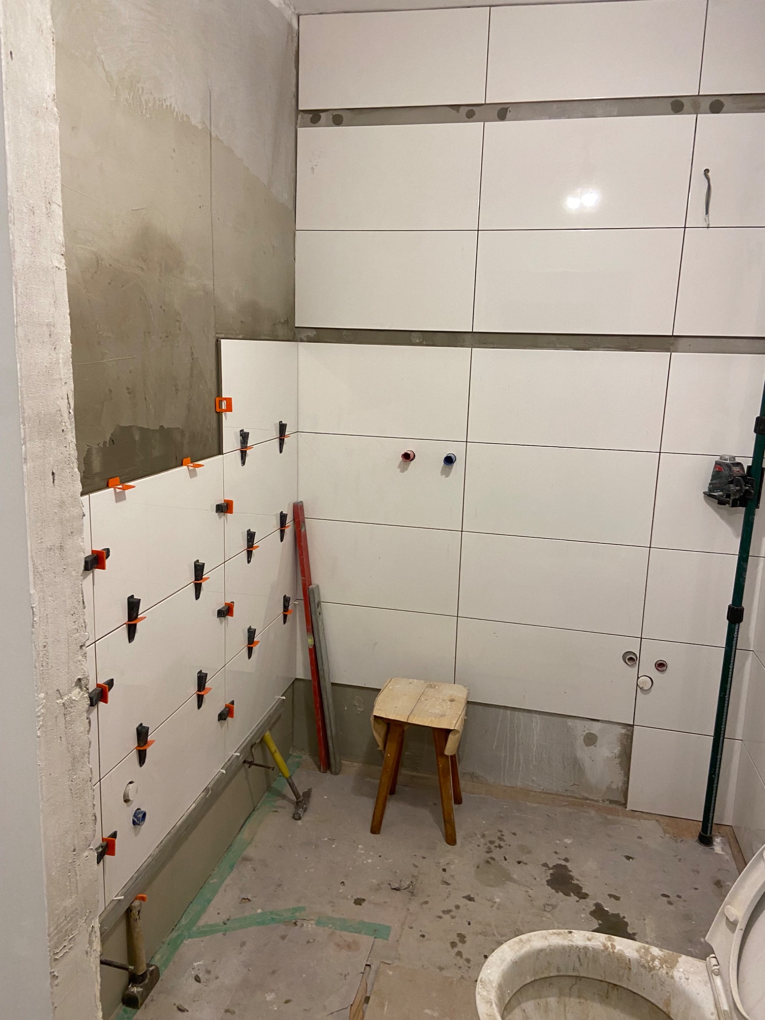 Białe płytki ścienne z pomarańczowymi i czarnymi klipsami poziomującymi w trakcie układania w łazience; widoczny fragment toalety, taboret, poziomica laserowa, młotek i fragment ściany bez płytek.