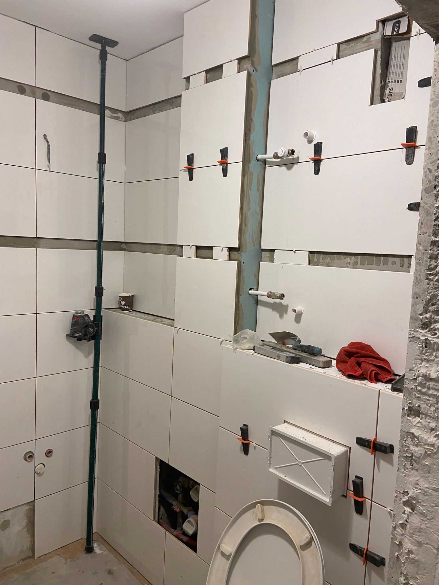 Montaż białych płytek w łazience, widoczny laser krzyżowy, narzędzia glazurnicze, elementy instalacji wodnej i fragment toalety w trakcie prac.