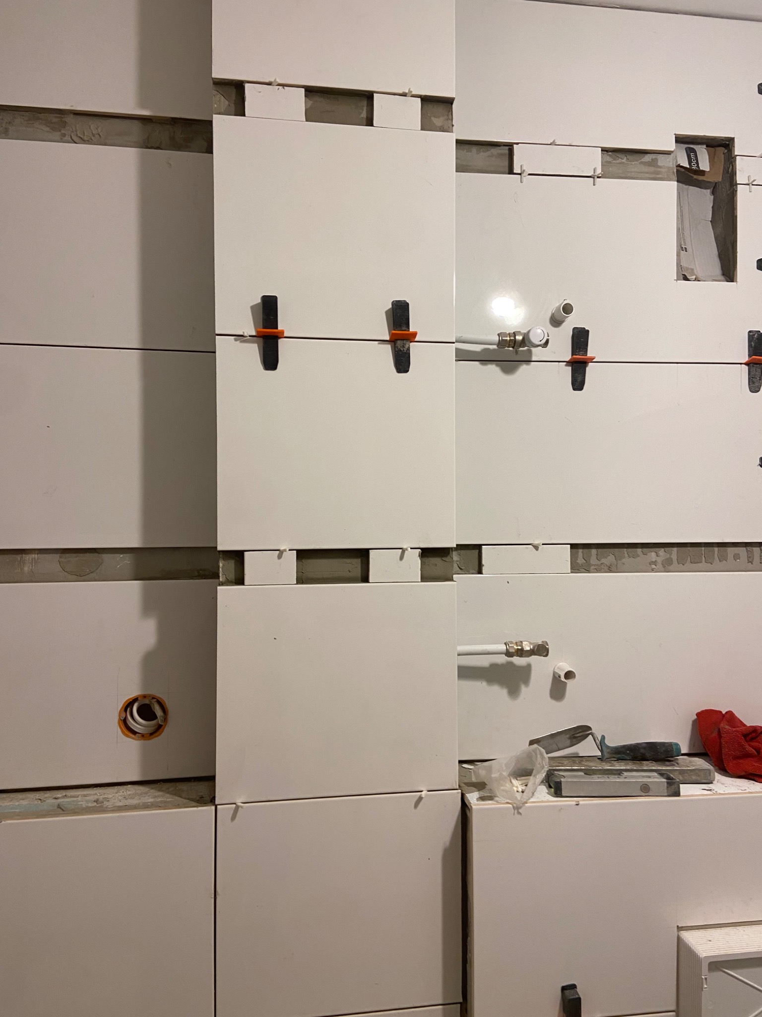 Układanie białych płytek ściennych z użyciem klipsów poziomujących; widoczne fragmenty instalacji wodnej i elektrycznej oraz narzędzia glazurnicze.