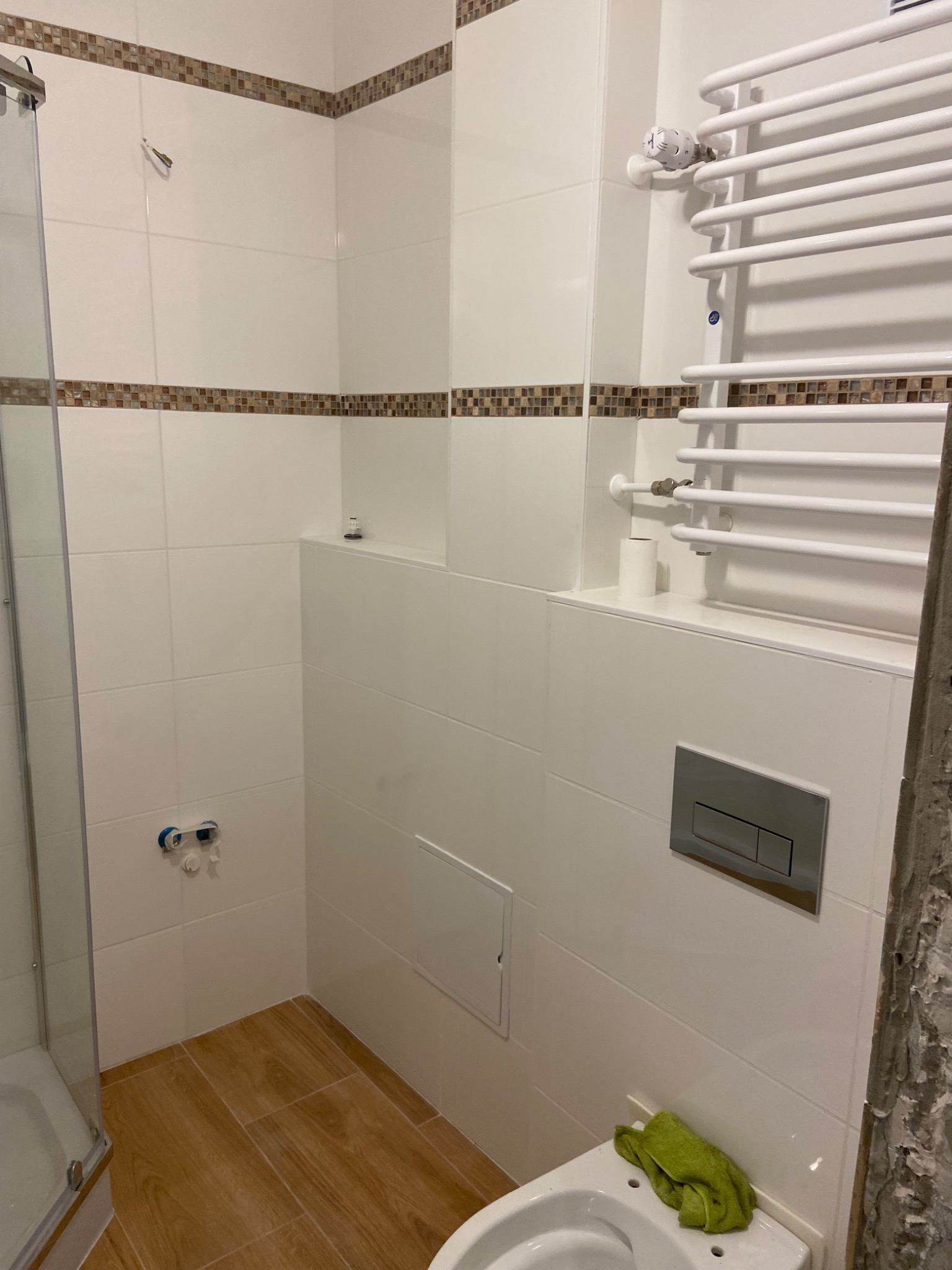 Wykończona łazienka z białymi płytkami, brązowym dekoracyjnym pasem, drewnopodobną podłogą, grzejnikiem drabinkowym i toaletą z zielonym ręcznikiem.