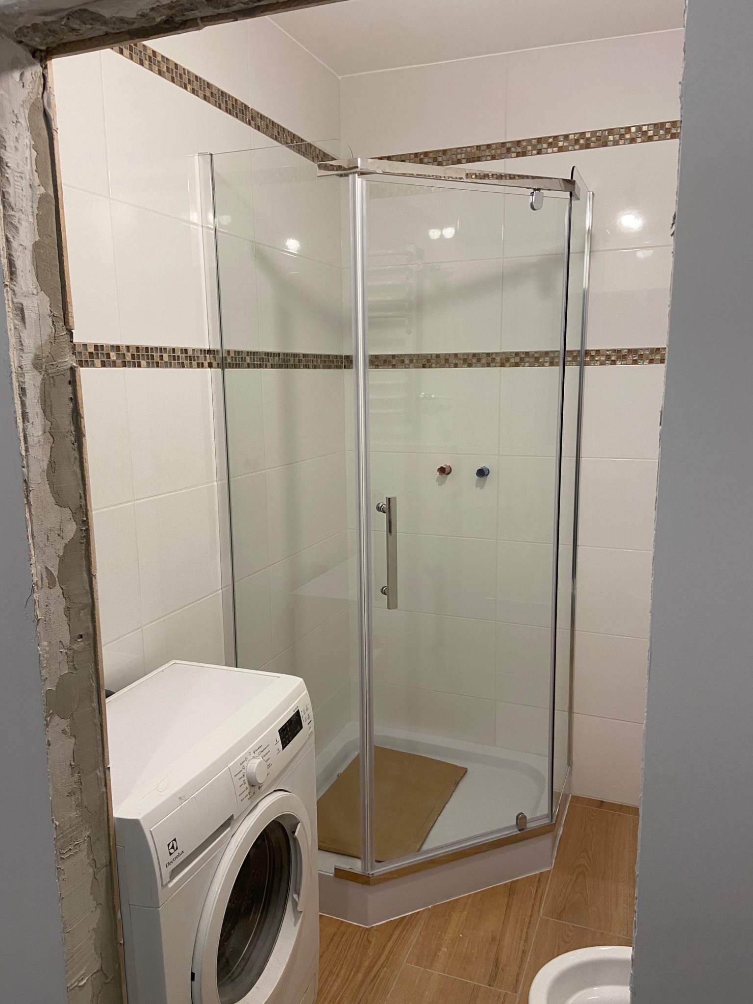 Narożna kabina prysznicowa z chromowanymi profilami i szklanymi drzwiami w łazience z białymi płytkami i dekoracyjnym pasem mozaiki, obok pralka Electrolux i fragment surowej ściany po remoncie.
