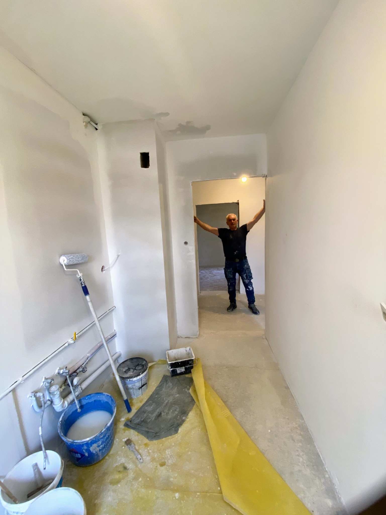 Wnętrze korytarza w trakcie prac wykończeniowych; widoczne ślady szpachlowania na ścianach, wiadro z farbą, wałek malarski oraz mężczyzna w oddali, opierający się o futrynę.