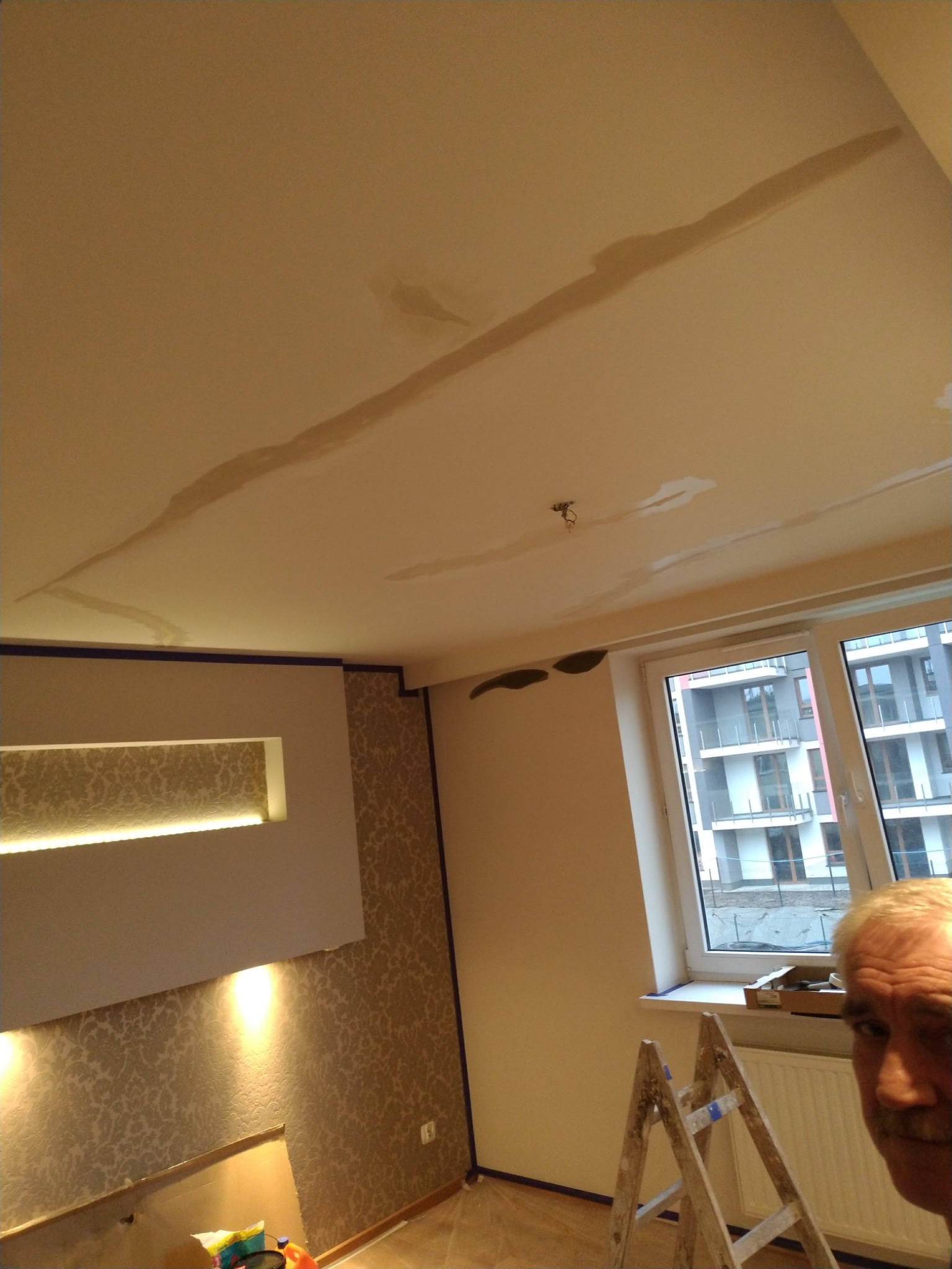 Pokój w trakcie prac wykończeniowych: widoczne ślady po szpachlowaniu na suficie, drabina oparta o ścianę przy oknie, fragment twarzy osoby w prawym dolnym rogu kadru.