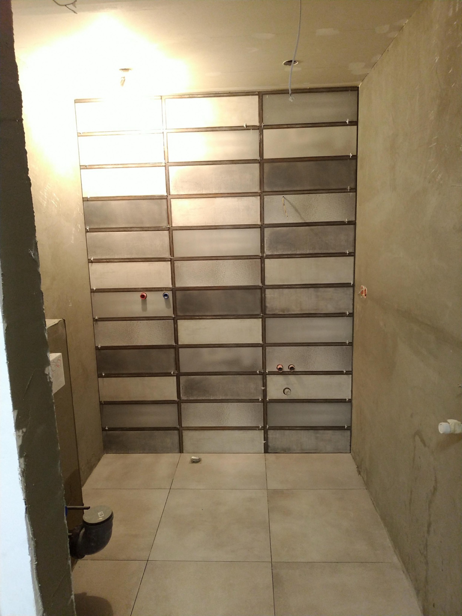 Wnętrze łazienki w trakcie prac w Krakowie: ułożone poziomo, prostokątne płytki w odcieniach szarości i beżu z widocznymi krzyżykami dystansowymi, surowa ściana z instalacją hydrauliczną...