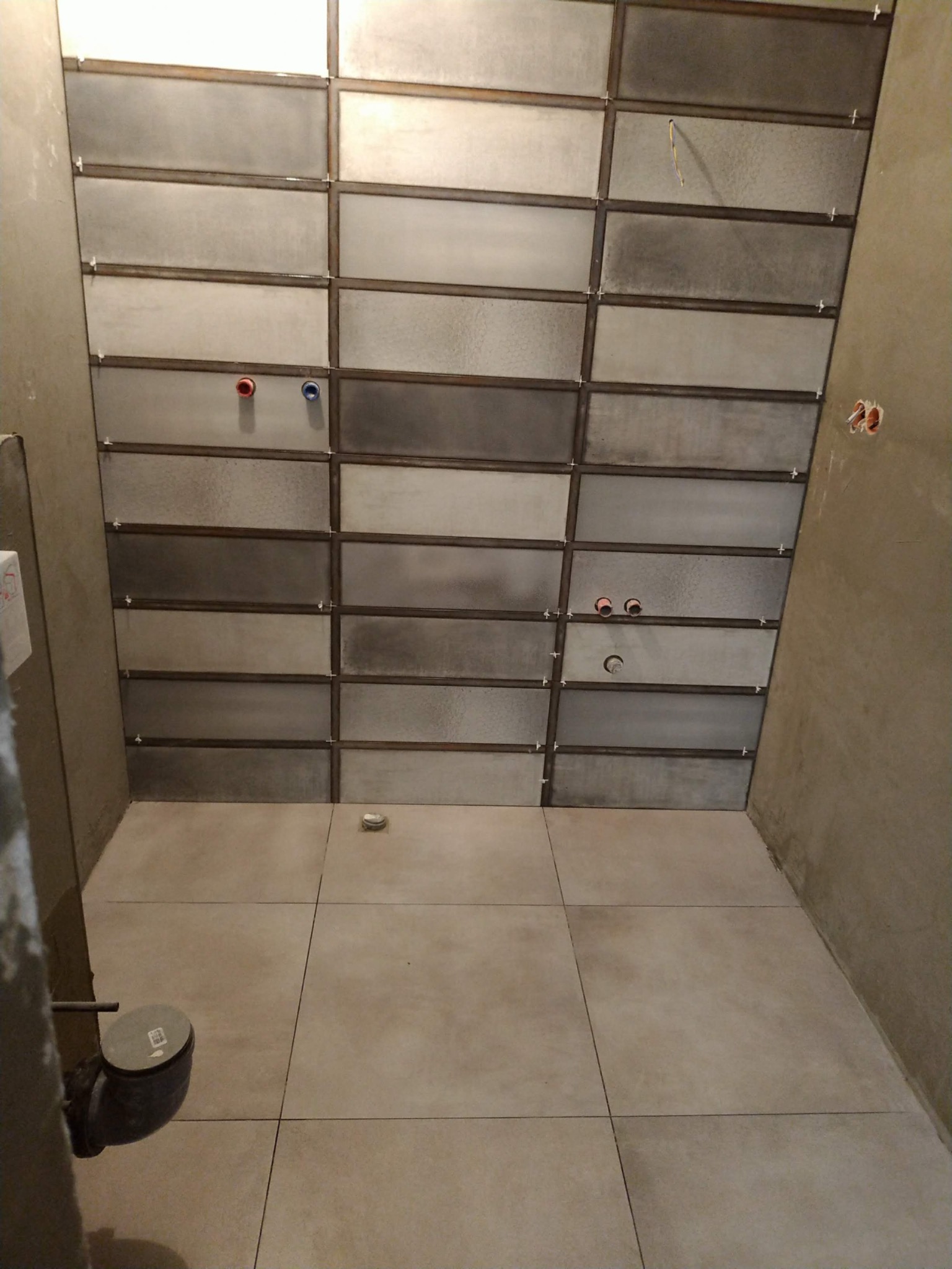 Wnętrze łazienki w trakcie prac: ułożone szare, prostokątne płytki na ścianie z widocznymi otworami na instalacje i krzyżykami dystansowymi, podłoga wyłożona jasnymi płytkami, surowy tynk...