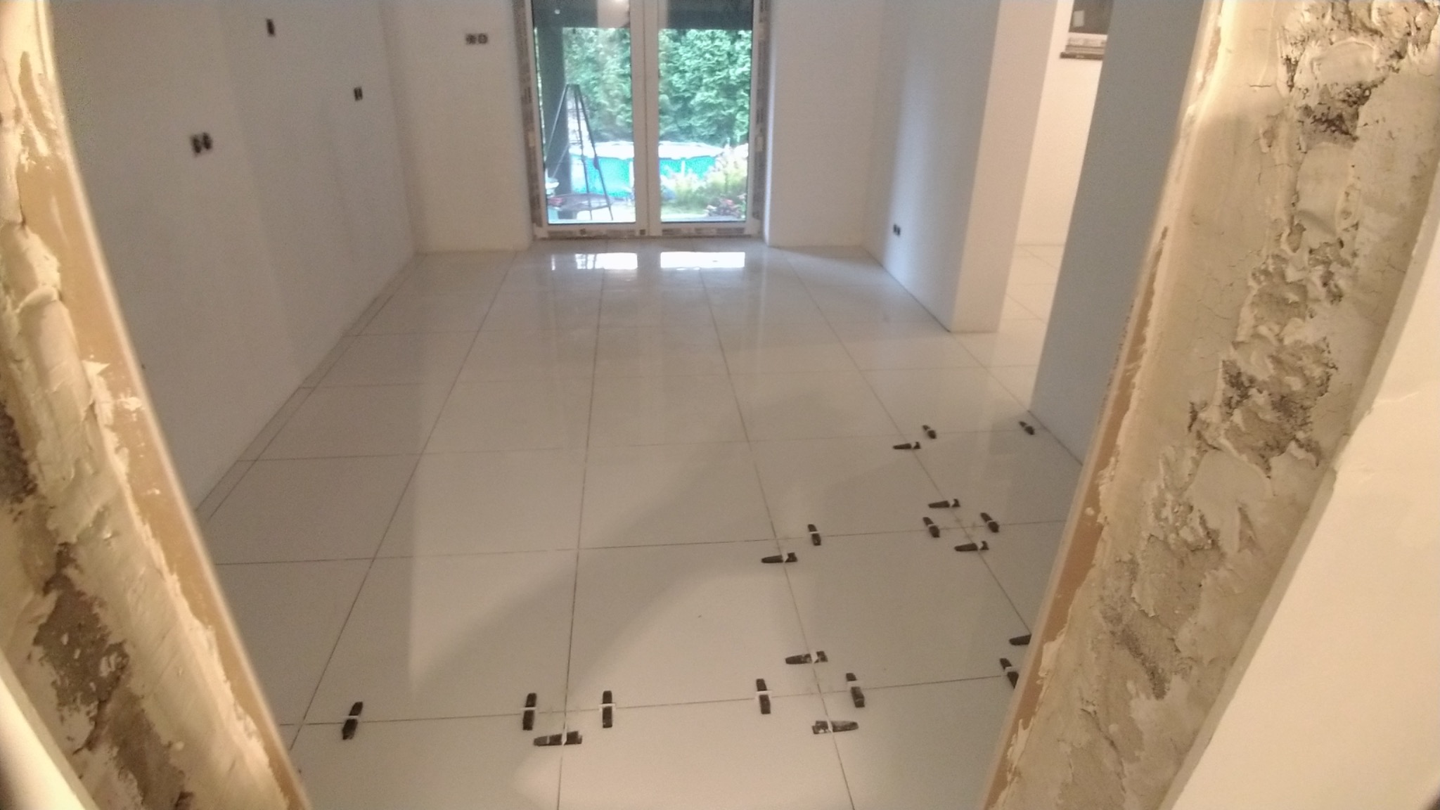 Białe płytki podłogowe w trakcie układania, widoczne plastikowe krzyżyki dystansowe, surowe ościeżnice drzwiowe z resztkami zaprawy, w tle okno balkonowe i drabina.