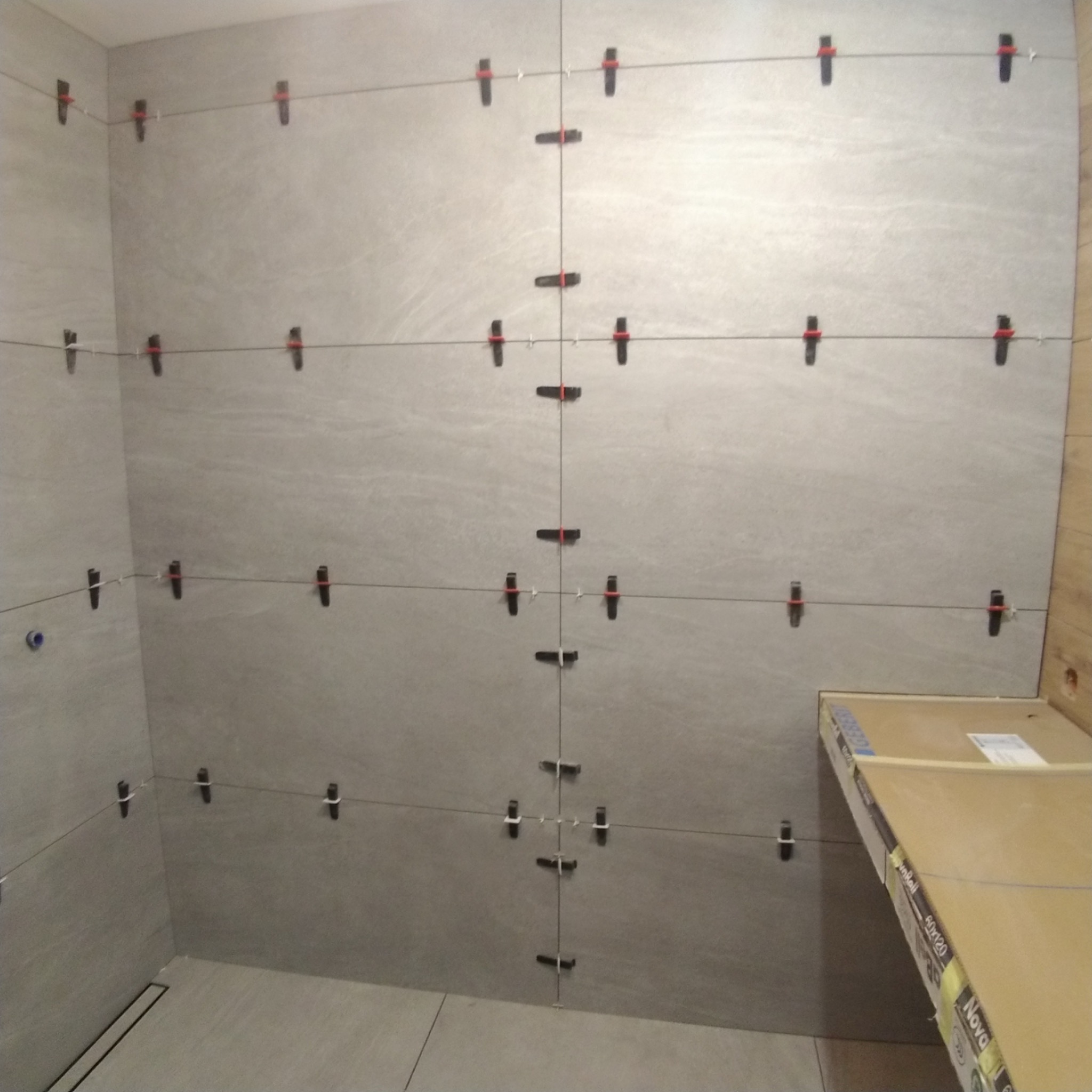 Ściana łazienki w trakcie układania szarych płytek wielkoformatowych, widoczne klipsy i sznurki poziomujące, w tle karton z płytkami.