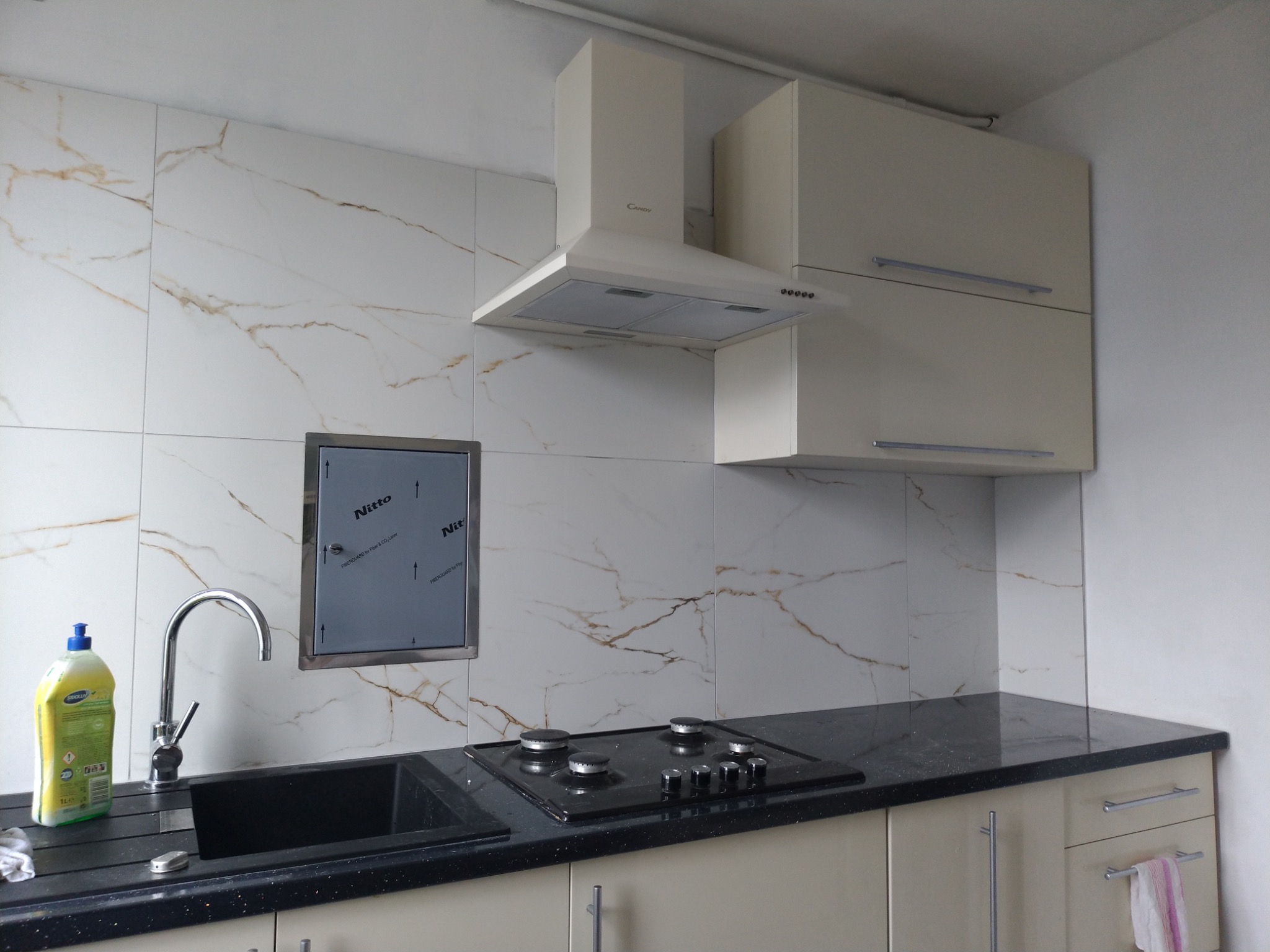 Wykończona kuchnia z białymi płytkami imitującymi marmur, czarnym zlewem i płytą grzewczą, beżowymi szafkami oraz okapem Candy.