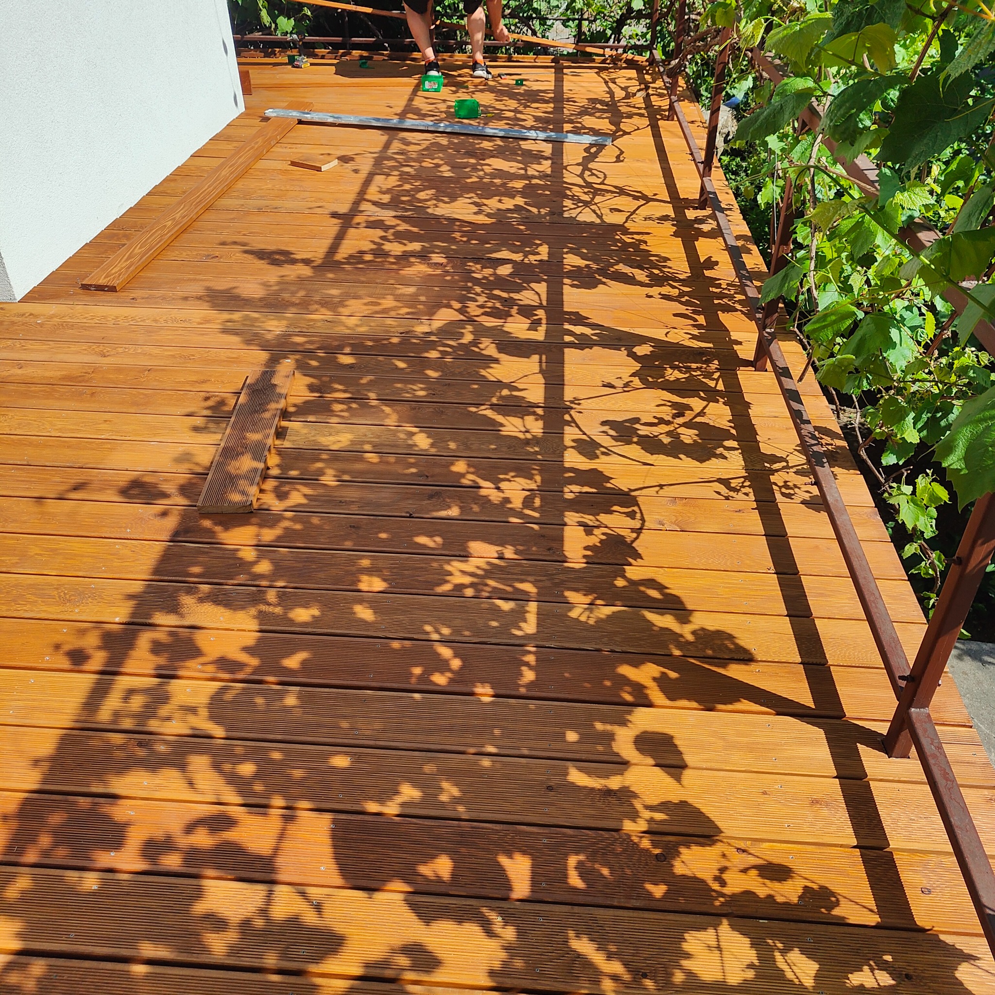 Drewniany taras w trakcie budowy, widoczne deski, elementy konstrukcyjne, cień liści i częściowo sylwetka osoby pracującej.