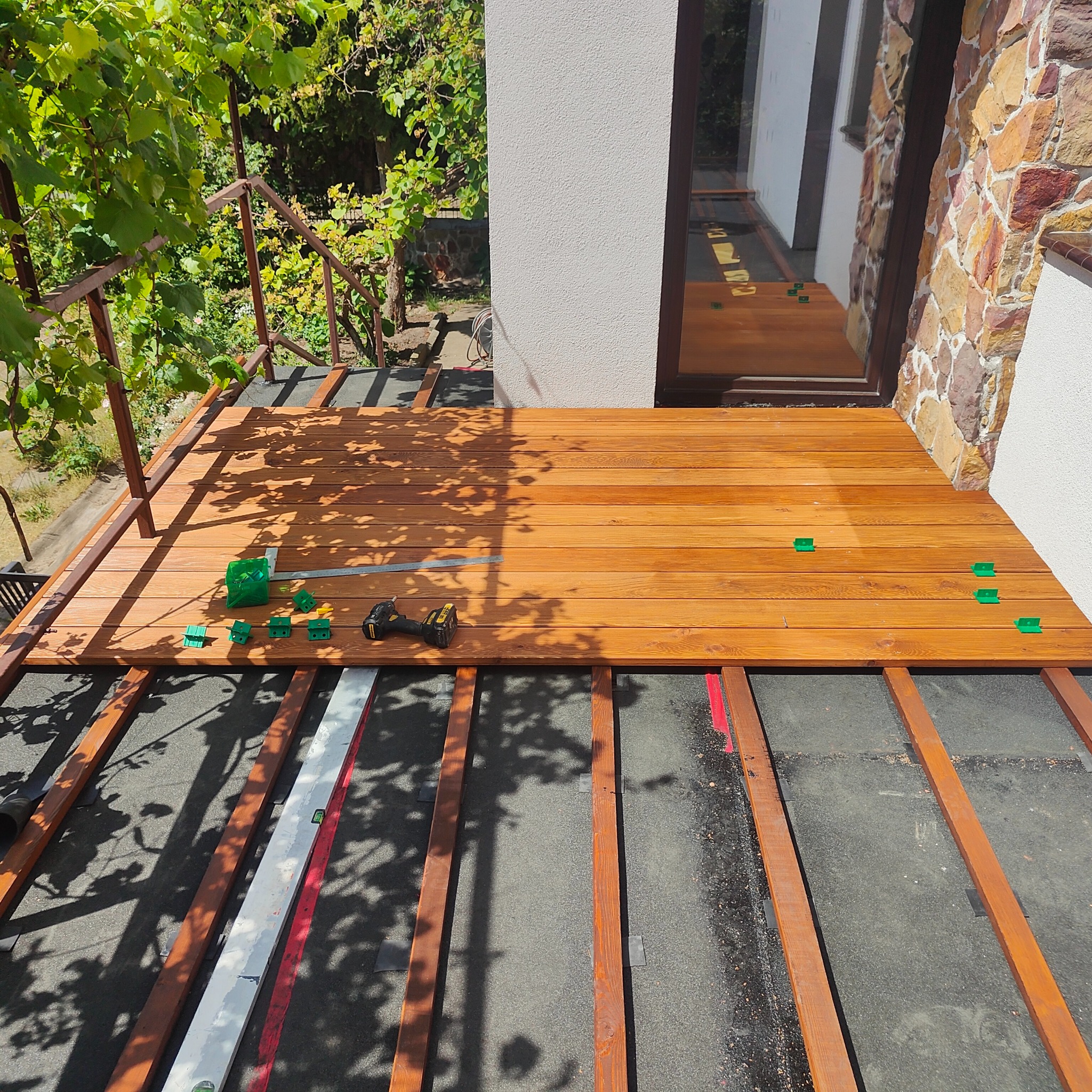 Montaż desek tarasowych na legarach z użyciem poziomicy i zielonych podkładek dystansowych, w tle widoczne pnącze winorośli.