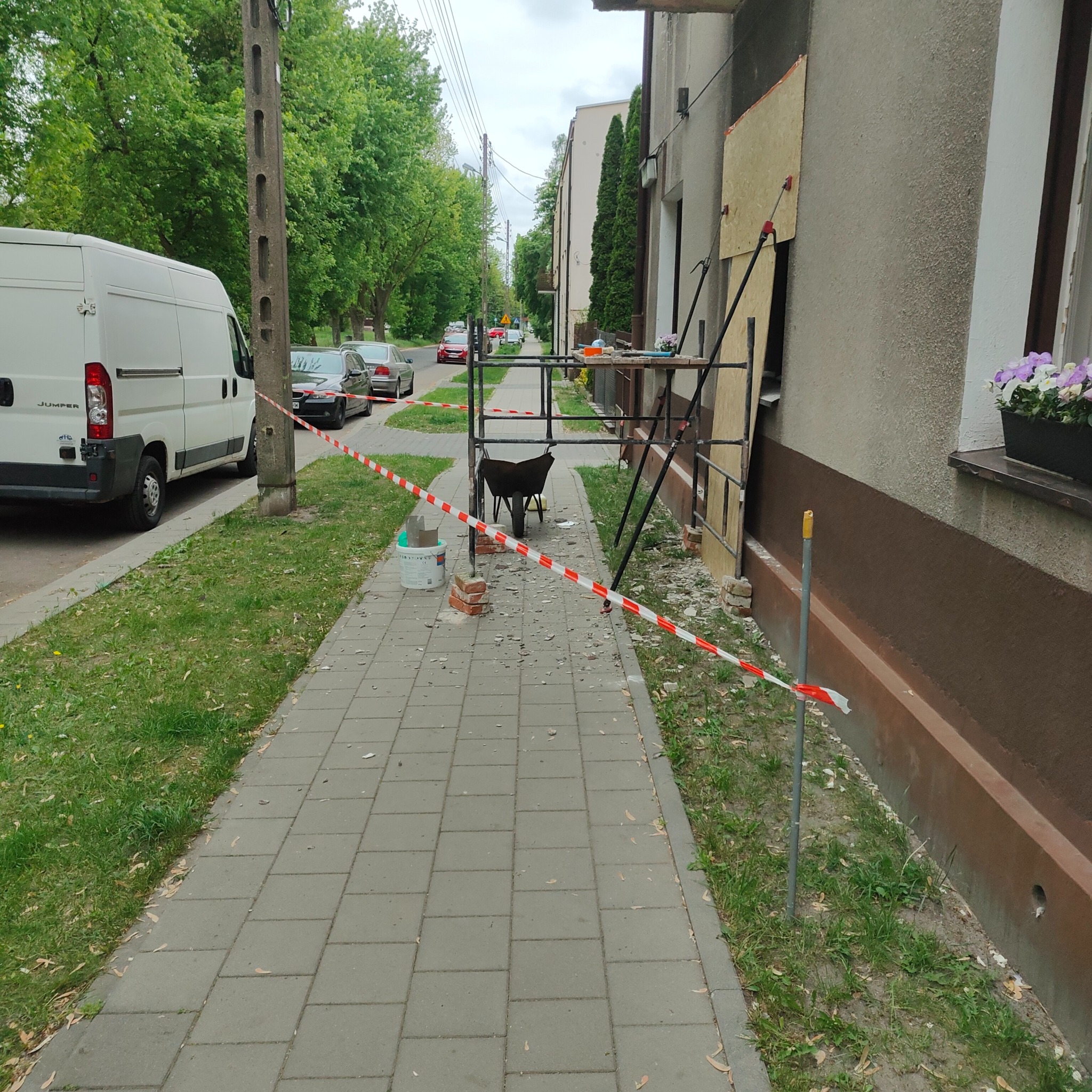 Przygotowania do remontu elewacji domu: rusztowanie, gruz, taśma ostrzegawcza, wiadro z farbą i taczka na chodniku, biały bus w tle.