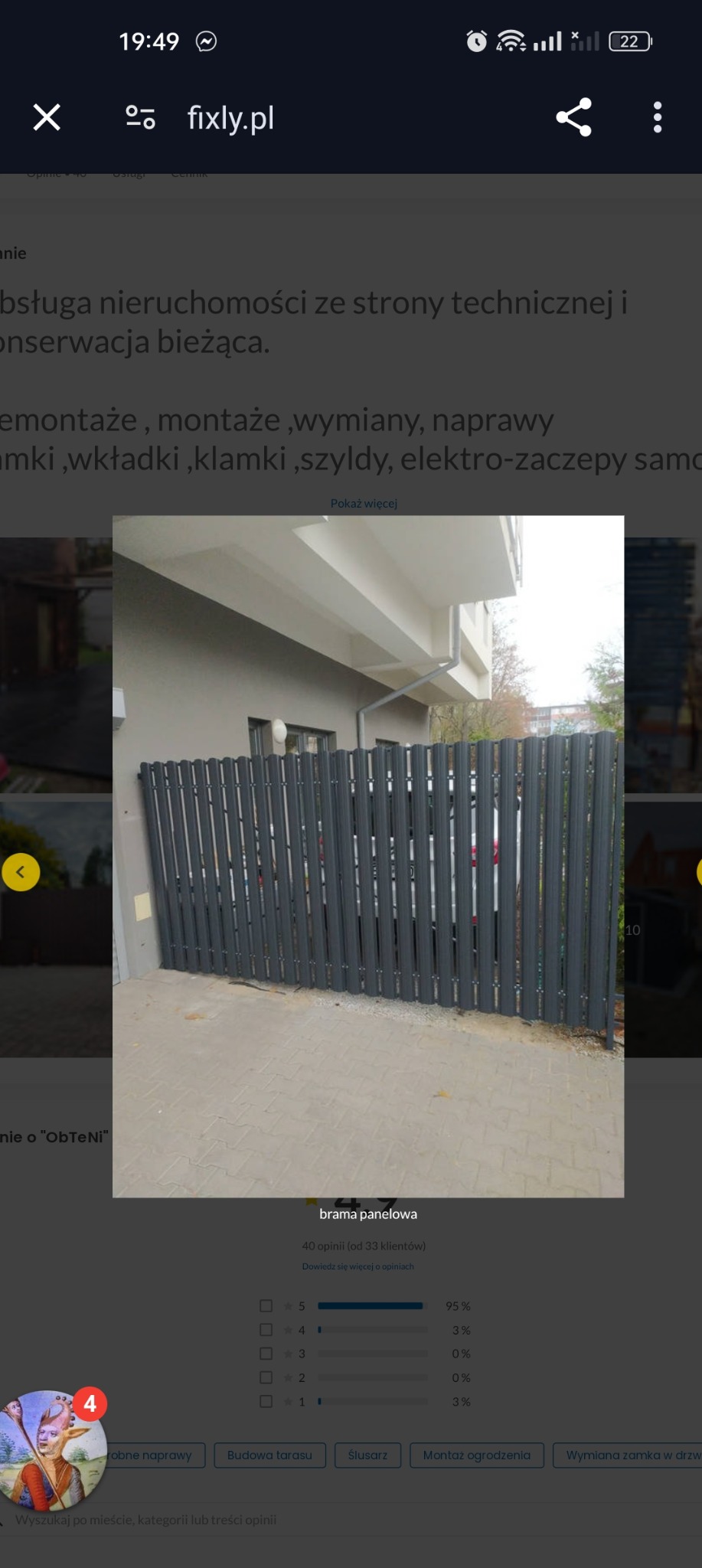 Szare ogrodzenie panelowe z pionowych, wąskich profili, zamontowane przed budynkiem z szarą elewacją i białym podcieniem, widok z poziomu chodnika z szarej kostki brukowej.