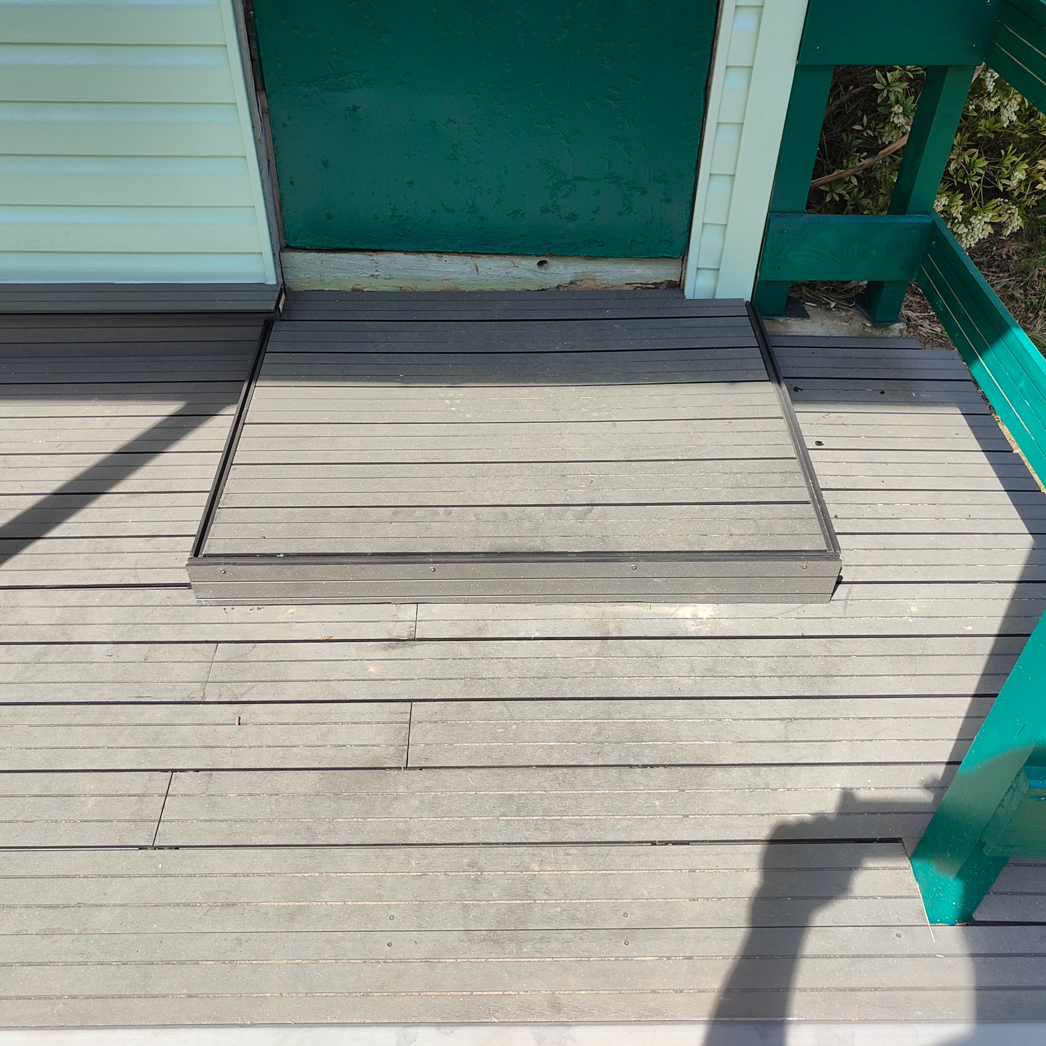Szary taras z desek kompozytowych z ukrytą klapą włazową, prowadzącą do piwnicy lub innego pomieszczenia pod tarasem, z fragmentem zielonego wejścia do budynku.