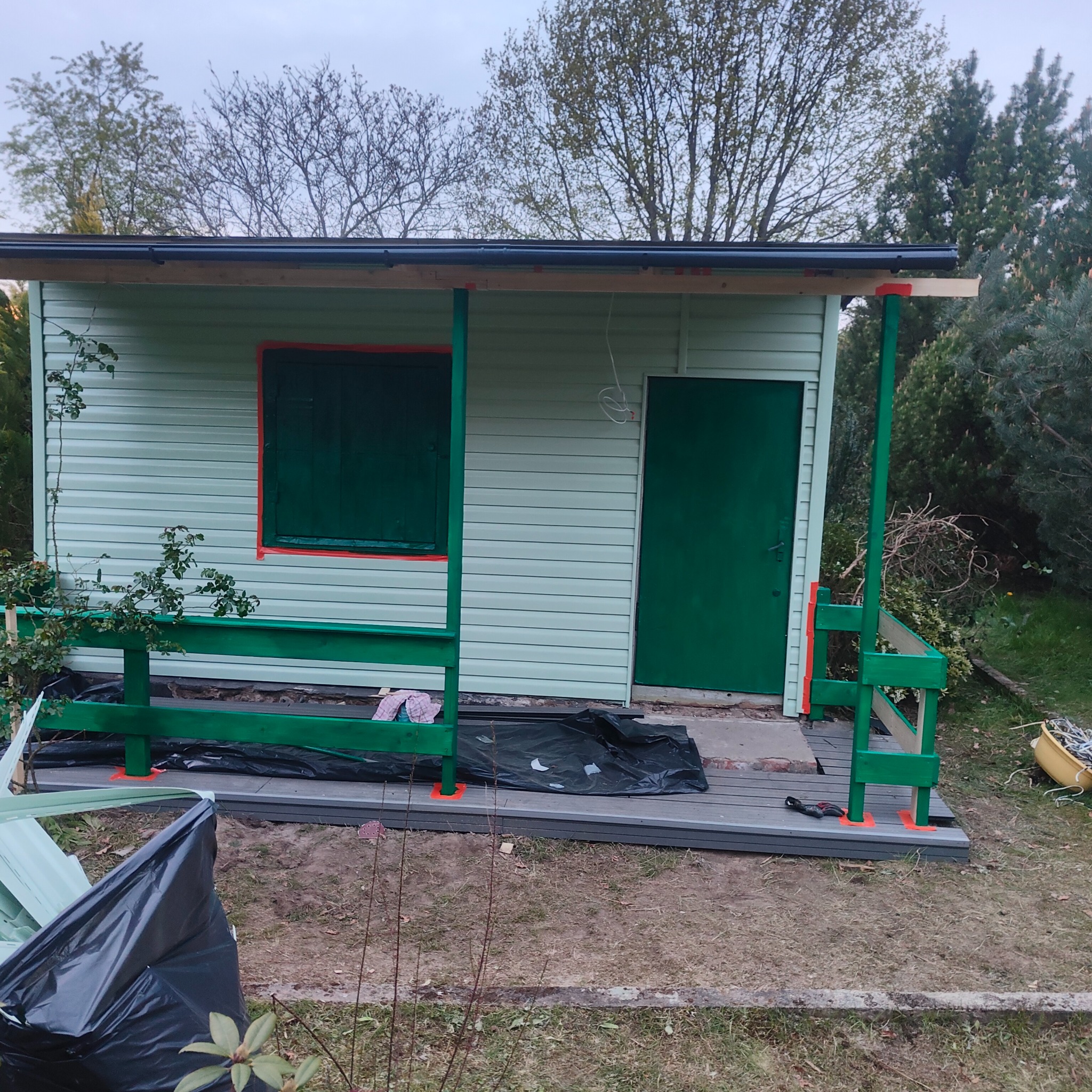 Niedokończona budowa małego domku z elementami tarasu kompozytowego, widoczne deski tarasowe, zielone elementy konstrukcyjne i worki z odpadami budowlanymi.